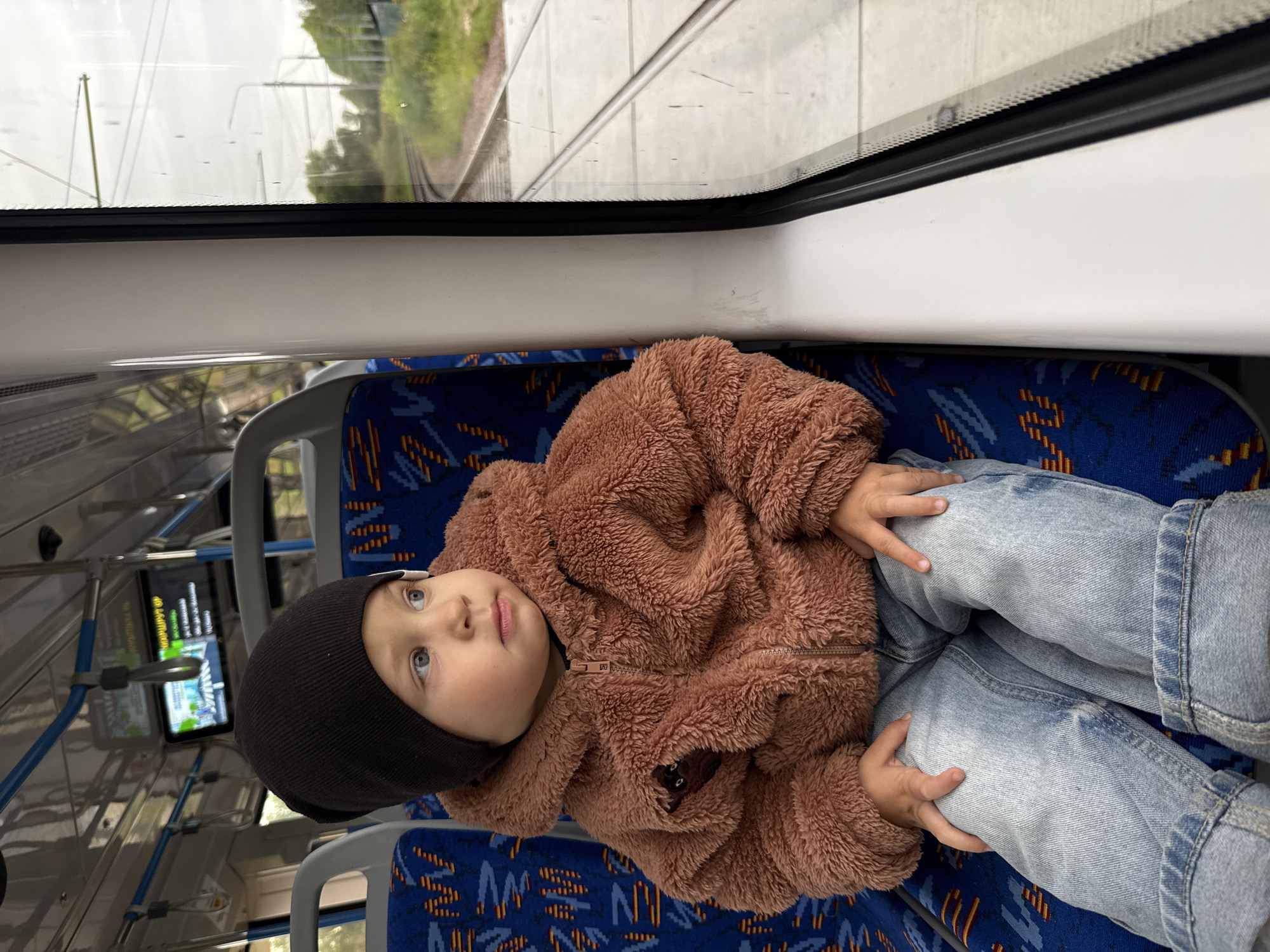 A young child, wearing a black beanie and a fuzzy brown jacket, sits on a blue and yellow patterned seat inside a public transport vehicle, likely a tram, as indicated by the tracks visible through the window. The child is looking out the window at the passing scenery of greenery and trees, with overcast skies suggesting a cool day. The interior of the vehicle shows other empty seats and overhead fixtures. The scene is set in Rusanovo, Russia.