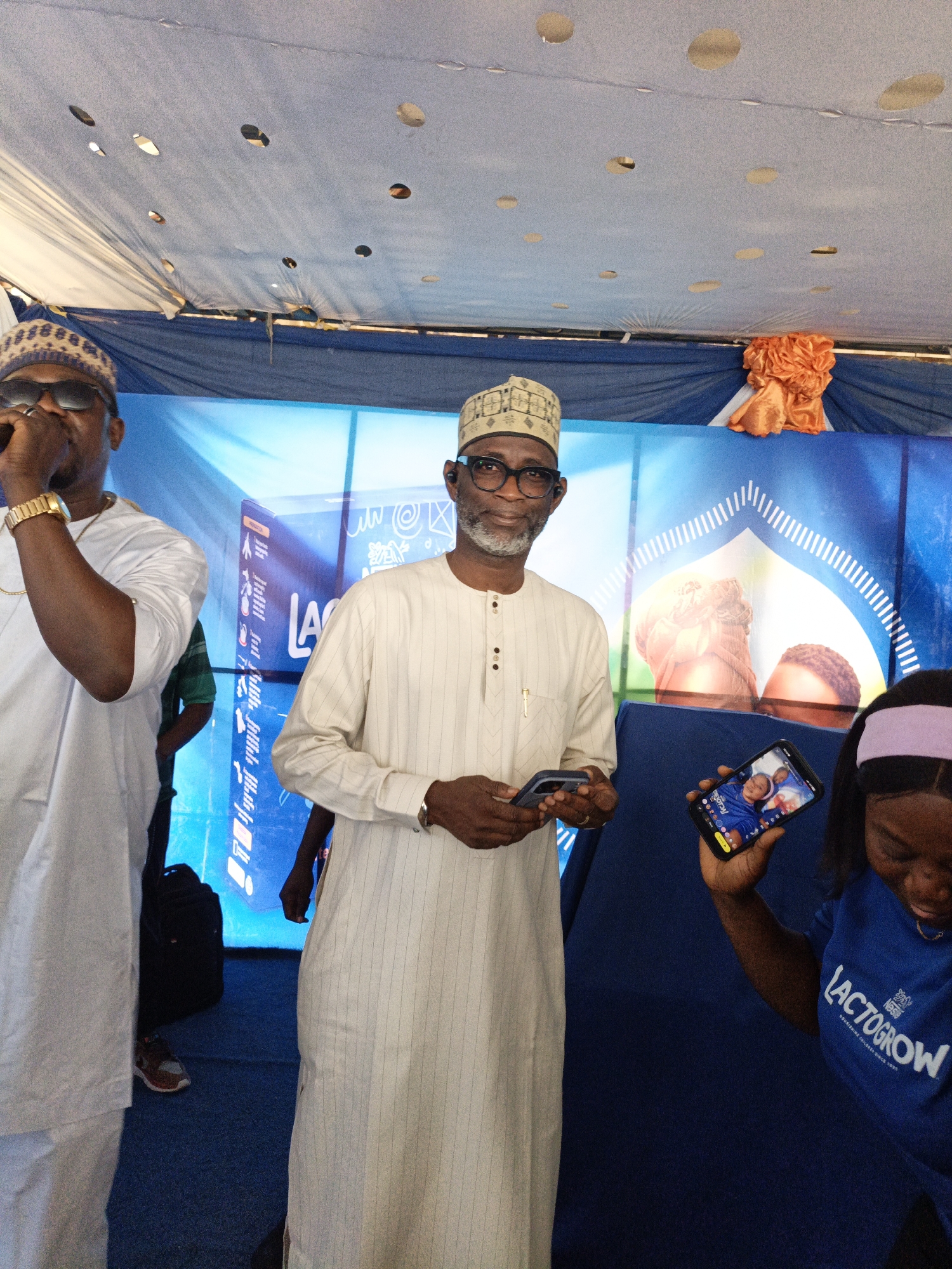 This media file captures an indoor event, likely a promotional gathering under a tent or temporary structure, characterized by a light blue fabric ceiling with circular cutouts. The scene is dominated by a vibrant blue color scheme, including a large blue backdrop and a dark blue carpeted floor.
Three individuals are prominent in the frame. On the left, a man dressed in white traditional attire with a patterned skullcap and dark sunglasses holds a microphone to his mouth, appearing to be speaking or performing. He wears a gold watch and chain. In the center, another man, wearing a white striped traditional tunic (agbada or similar) and a patterned skullcap, stands smiling directly at the camera. He wears glasses and holds a smartphone in both hands. Partially visible on the right, a woman wears a blue t-shirt that clearly displays the text "NESTLÉ LACTOGROW" and "NOURISHING CHILDREN SINCE 1866". She also wears a light purple headband and is focused on a smartphone in her hand, which shows an image of a child's face and the "LACTOGROW" logo.
The blue backdrop behind the subjects features the brand name "LACTOGROW" prominently in white text, alongside various graphics including product packaging, an arched design, and what appears to be abstract patterns and possibly Arabic script. An orange decorative fabric element is visible at the top right of the backdrop.
The event appears to be a daytime gathering, given the bright lighting. The traditional attire of the men suggests a cultural context, fitting with the specified location of Tarauni, Nigeria. The overall mood conveyed by the central man's smile is positive and engaging.