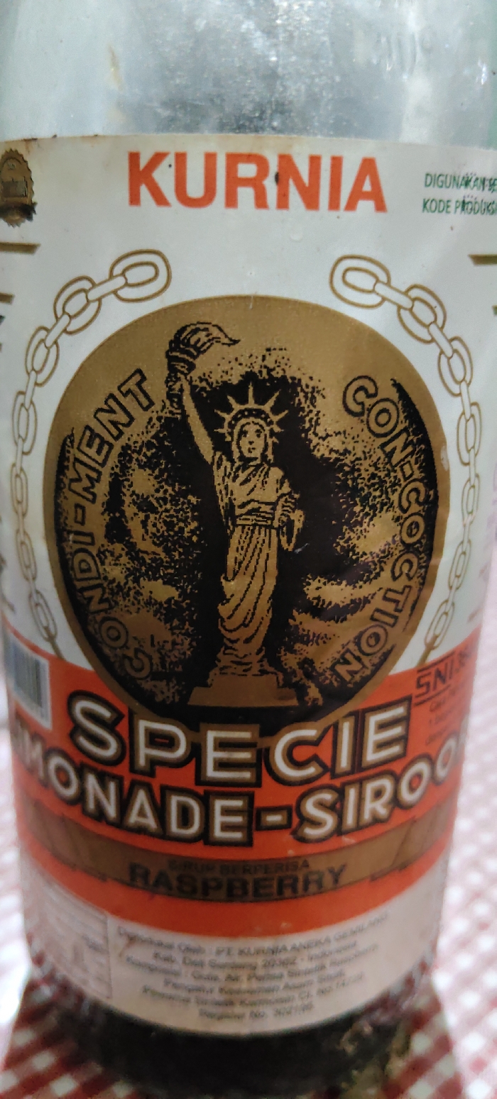 A clear glass bottle, partially out of frame, rests on a red and white checkered tablecloth. The bottle displays a white and orange paper label featuring the brand name "KURNIA" in orange capital letters at the top. Centrally, a circular gold and black emblem depicts a stylized Statue of Liberty holding a torch, encircled by the text "CONDIMENT CONFECTION" and a gold chain pattern. Below this, "SPECIE LEMONADE-SIROOP" is printed, with "RASPBERRY" identified as the flavor. The lower portion of the label contains indistinct Indonesian text, which includes "Diproduksi Oleh PT KURNIA ANEKA GEMILANG, Kab. Deli Serdang 20362 - Indonesia," indicating the manufacturing location, and an SNI certification mark. The upper section of the bottle is slightly out of focus. The scene is located in Dayah Tuha, Indonesia.