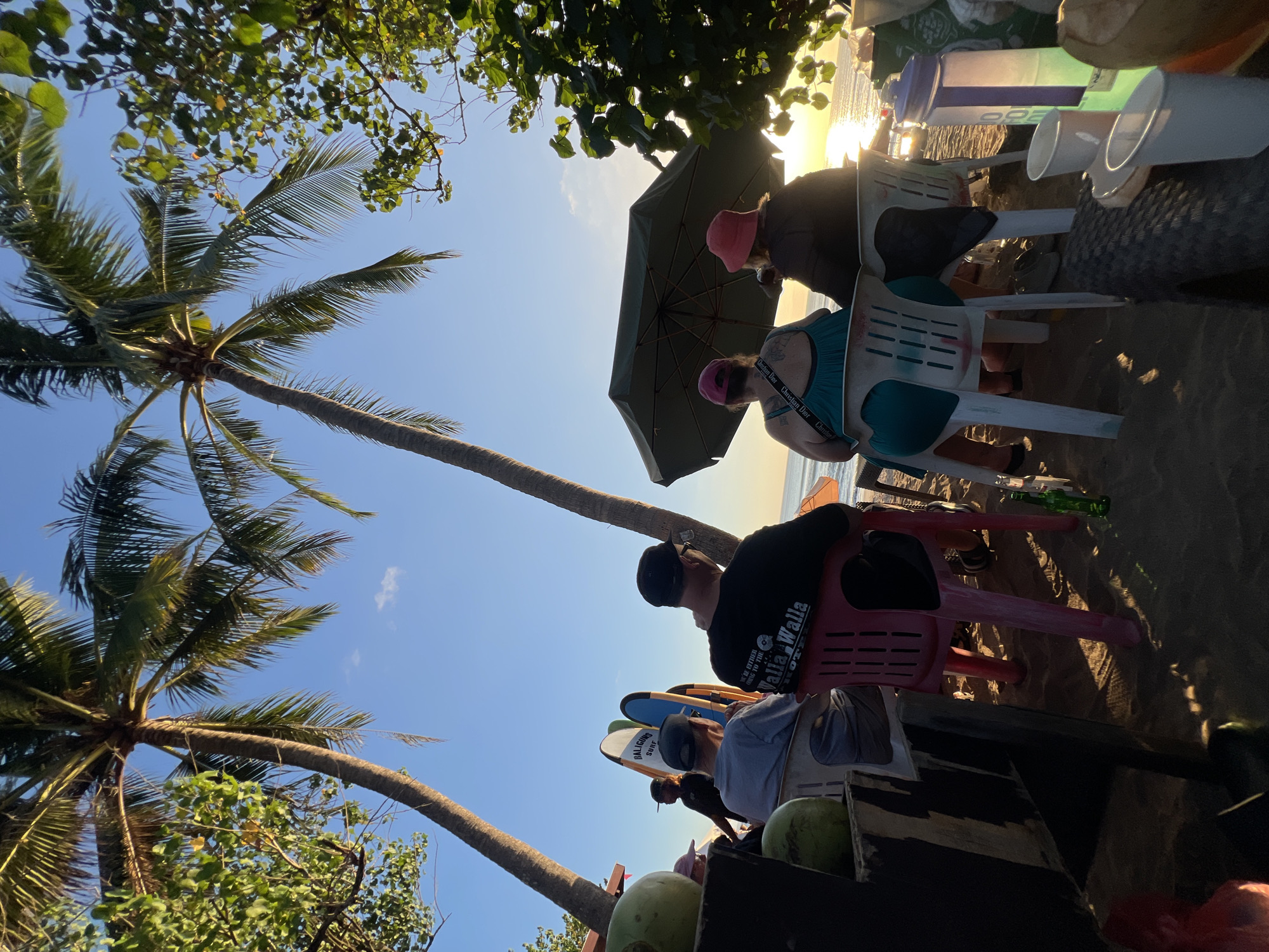 The image captures a tranquil beach scene in Kuta, Indonesia, during the golden hour. Several people are seated on plastic chairs on the sand, facing the ocean. Two individuals are under a large umbrella, their backs to the camera, enjoying the sunset. In the foreground, there are surfboards propped up, suggesting a surfing community. Coconut trees frame the scene, their fronds gently swaying under the clear blue sky. The overall atmosphere is relaxed and serene, with the warm glow of the setting sun casting long shadows on the beach.
