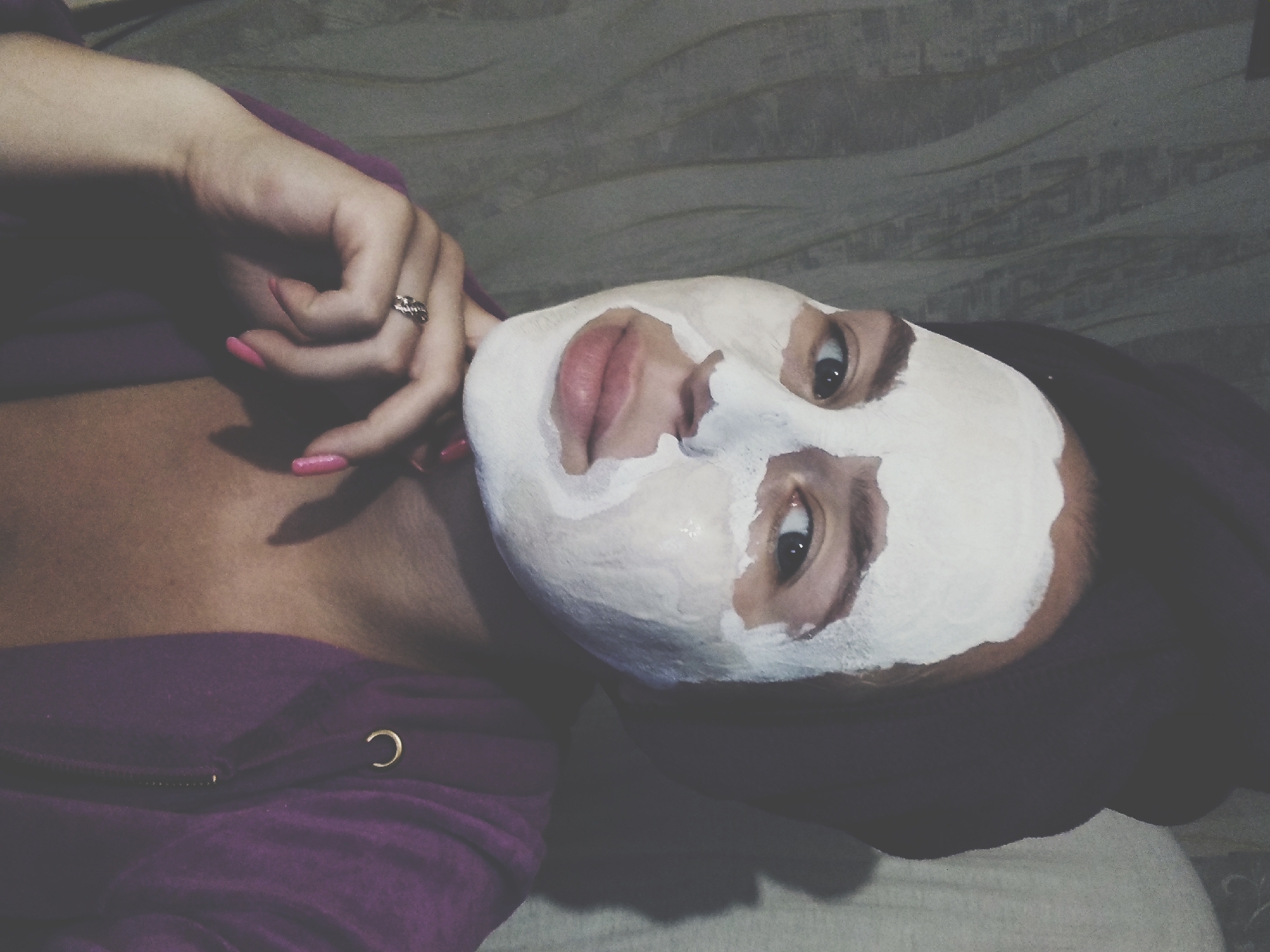 The image shows a person with a face mask on, lying down. The person is wearing a purple hoodie. The background appears to be a bed or sofa with a textured gray blanket. The lighting is dim, suggesting it might be evening or indoors. The person's hand, with bright pink nail polish and a ring, is resting near their face. The overall mood is relaxed and private, perhaps indicating a self-care moment. There is no visible text in the image. The location context provided is Radisne, Ukraine.