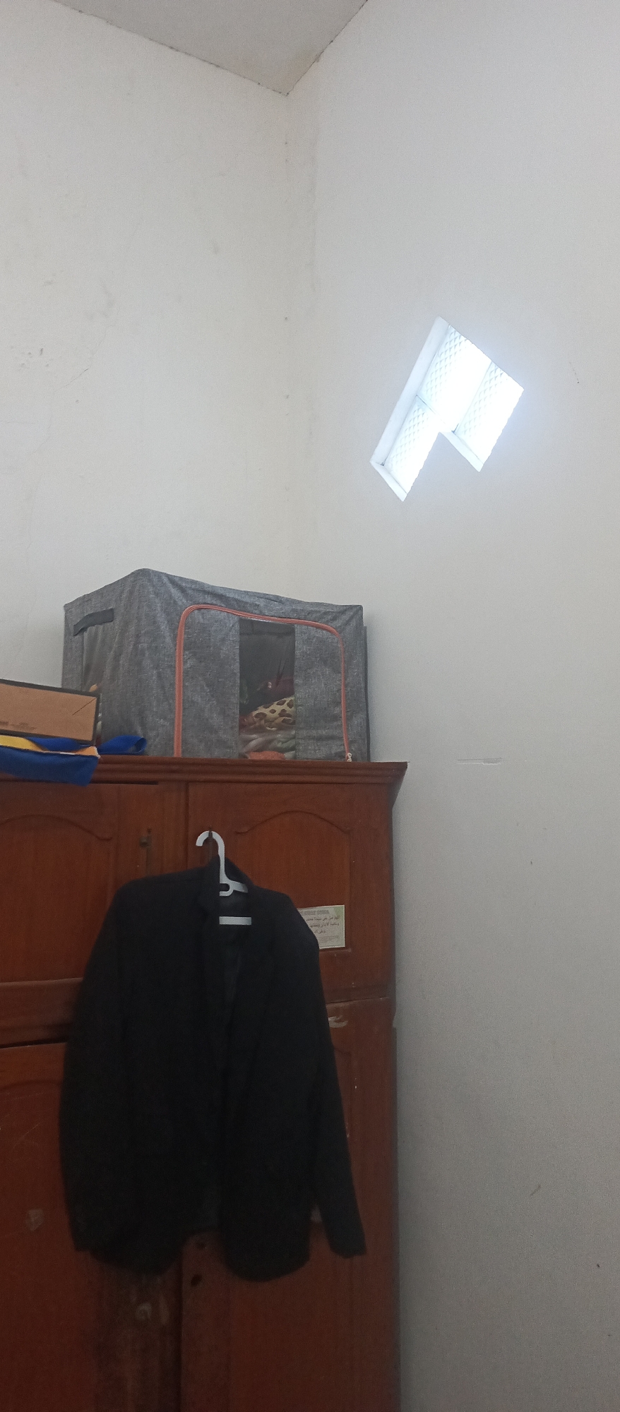 The image depicts a dark blazer hanging on a white plastic hanger on a wooden cabinet. The blazer is black. The cabinet appears to be made of dark wood and has two doors. Above the cabinet is a gray fabric storage bin with orange trim, partially open, revealing clothing inside. To the left of the storage bin, a stack of books is visible. The background is a white wall with a small, angled window near the ceiling, through which light is visible. The scene is indoors and appears to be in a room. There is a piece of paper with writing on it attached to the right door of the cabinet. The location is stated to be in the city of Arjasa, Indonesia.