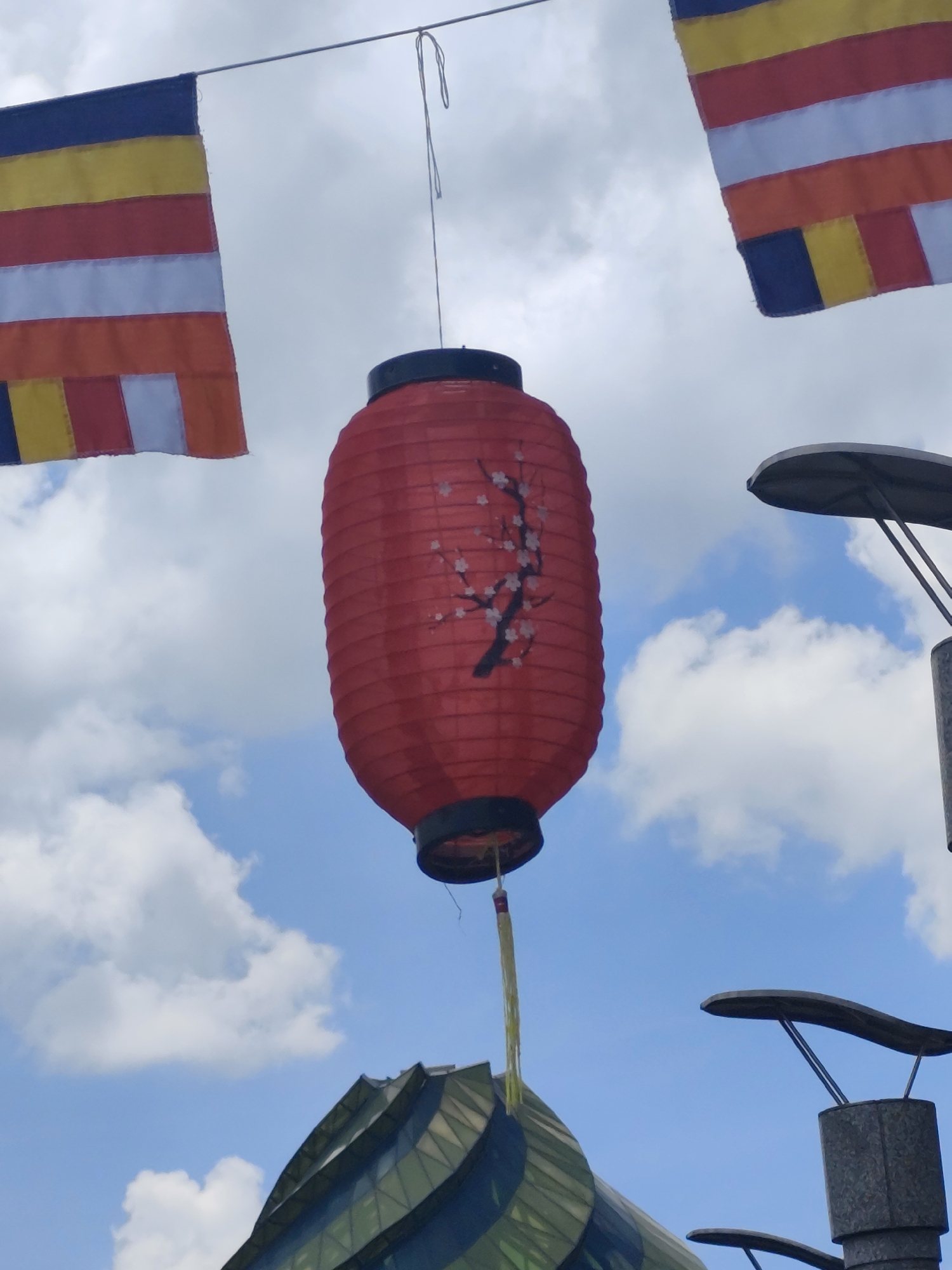 The image shows a red, barrel-shaped lantern with a black top and bottom, suspended by a string. The lantern is decorated with a dark-colored, flowering branch design. Above the lantern, two flags with horizontal stripes of blue, yellow, red, white, and orange are visible against a backdrop of blue sky with white clouds. Below the lantern, part of a building with green-tinted glass is visible, along with a street lamp. The scene is located in Da Lat, Vietnam.