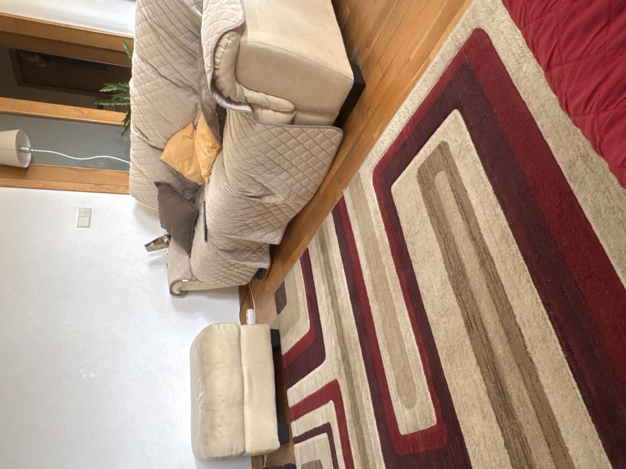 The image shows an interior setting with a beige, overstuffed sofa placed on a hardwood floor adjacent to a colorful rug with geometric patterns in red, brown, and beige. The rug partially extends under a red-colored object, likely a sofa or bedding. To the left of the sofa, against a white wall, stands a beige ottoman. A window with a lamp affixed to the wall is visible in the background. No people are visible in the image. The location, Mirandela, Portugal, cannot be confirmed from the visible details.