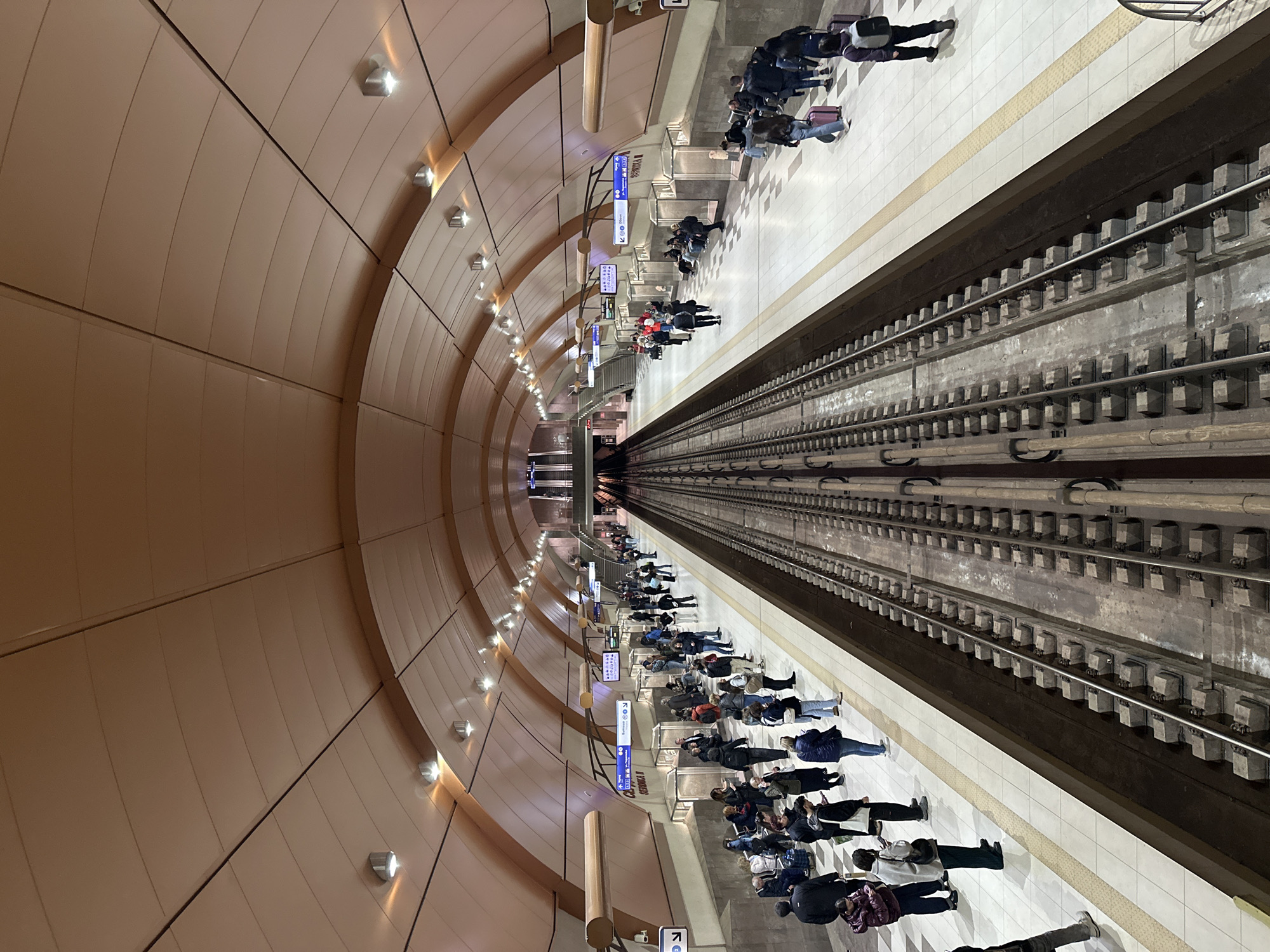 The image captures a long, modern subway station platform and tracks in Sofia, Bulgaria, viewed down the tunnel's length. The tracks run centrally into the distance, flanked by platforms on both sides.
Numerous people are congregated on the platforms, mostly standing or seated, patiently waiting for an arriving train. Many are dressed in jackets and coats, suggesting cool weather, and some are accompanied by wheeled luggage, indicating they are travelers or commuters.
The station's architecture is contemporary, featuring a high, uniformly arched ceiling composed of light brown or beige-colored panels. A series of metallic, upward-facing lights illuminate the ceiling with a warm glow, providing the primary light source for the station. The platforms are paved with light-colored square tiles, bordered by a textured warning strip adjacent to the tracks. The robust subway tracks, complete with concrete sleepers and metal rails, stretch far into the tunnel until they vanish into the receding darkness.
Above the platforms, several blue and white informational signs are visible. Clearly legible text includes "СЕРДИКА" (Serdika), a known metro station in Sofia, and phrases like "СПИРКА МЕТРО" (Metro Stop) and "СПИРКА БУС" (Bus Stop). These signs also display directional arrows and symbols for metro and bus connections. The overall scene depicts a busy, well-lit, and functional underground transit environment.