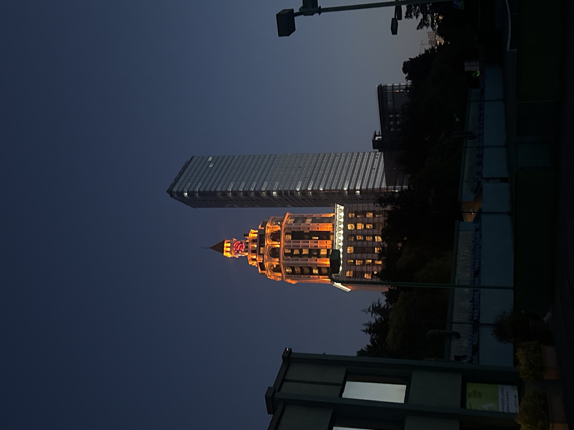 The image shows two buildings in Batumi, Georgia, at night. One building is a modern skyscraper with a reflective glass facade. The other is an ornate, tiered building with warm, glowing lights and a red "S" logo on its spire. The buildings are framed by trees, a street lamp, and parts of other structures, all set against a dark blue sky.