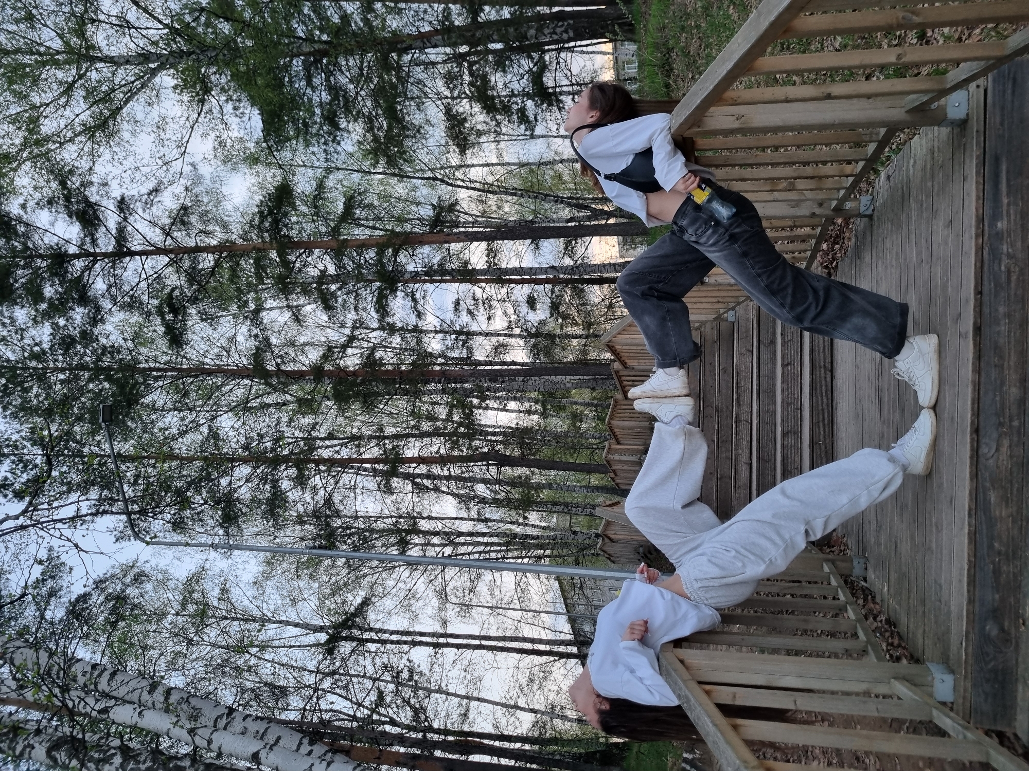 A long shot captures two young women on a wooden deck or stairs. The woman on the right wears a white cropped top with a black bralette, dark jeans, and white sneakers. She leans against a wooden railing, gazing upwards. The woman on the left wears a white sweatshirt and light gray sweatpants, also with white sneakers. She is similarly leaning back against a railing. The wooden structure is surrounded by tall, slender trees with sparse leaves. The perspective suggests the photo was taken from below, looking upwards through the trees. This scene is likely located within a park in the city of Chaikovsky, Russia.