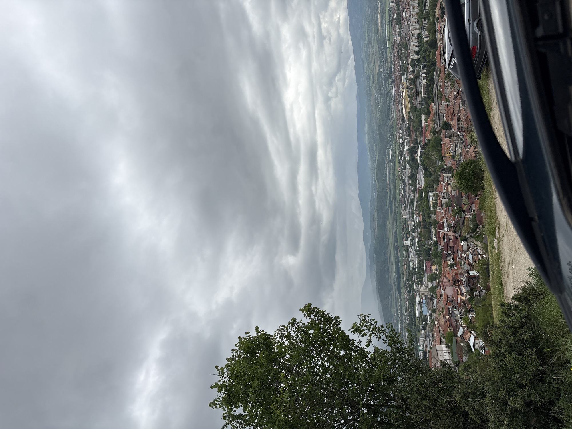 A high-angle, angled, long shot reveals a city from an elevated perspective, likely a vehicle. Overcast skies dominate the upper two-thirds of the image. The city buildings below are dense, with red rooftops and various architectural styles. A mountain range forms a backdrop to the city. A green tree is at the lower left corner, and the frame of a vehicle partially obscures the right side of the view.