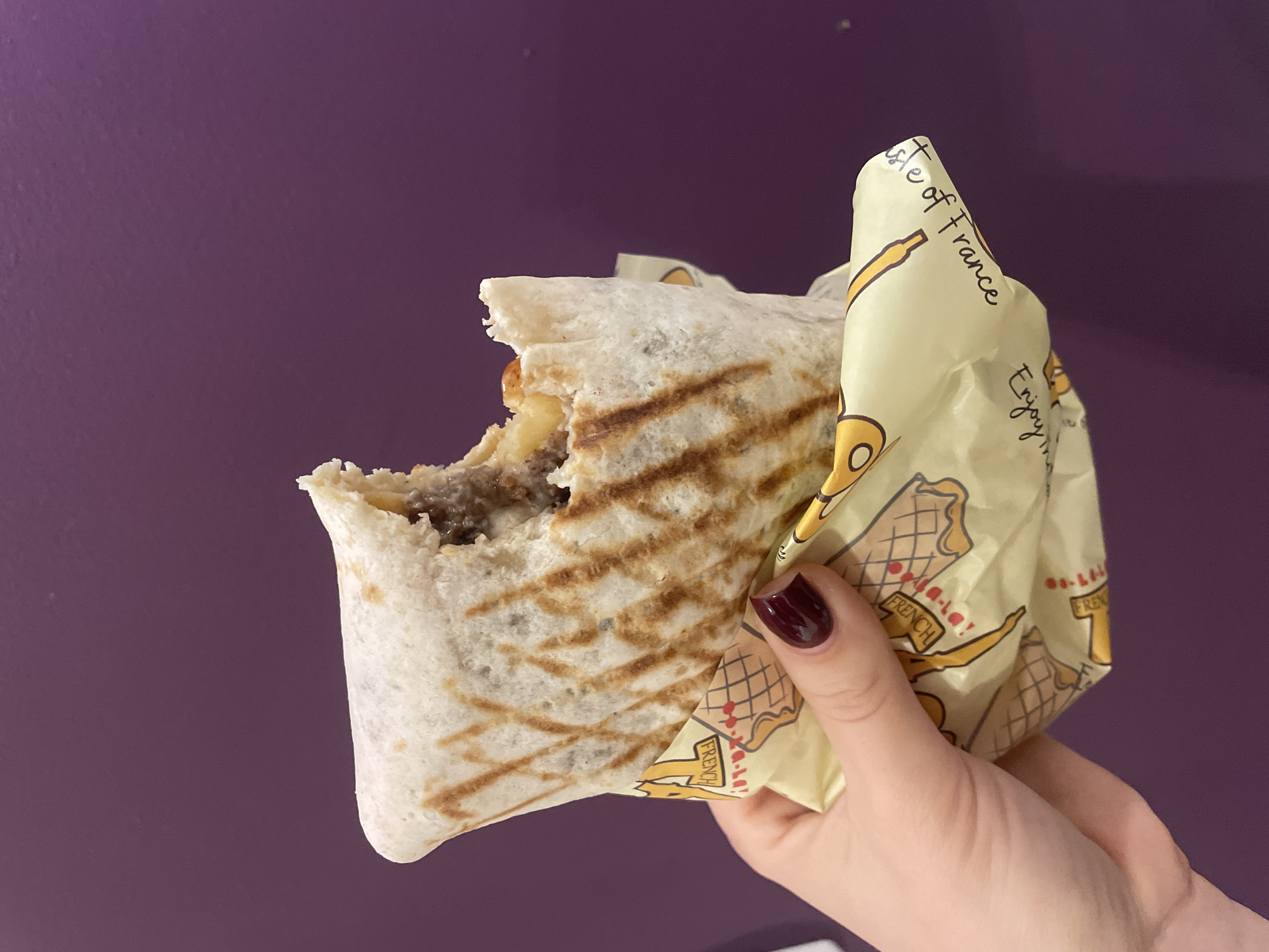 A hand holds a partially eaten wrap in its wrapper against a plain purple background. The wrap has grill marks and appears to contain ground meat and cheese. The wrapper is light yellow with a graphic depicting French pastries. The hand has manicured nails painted a dark red. The wrap does not seem to be in Sofia, Bulgaria.