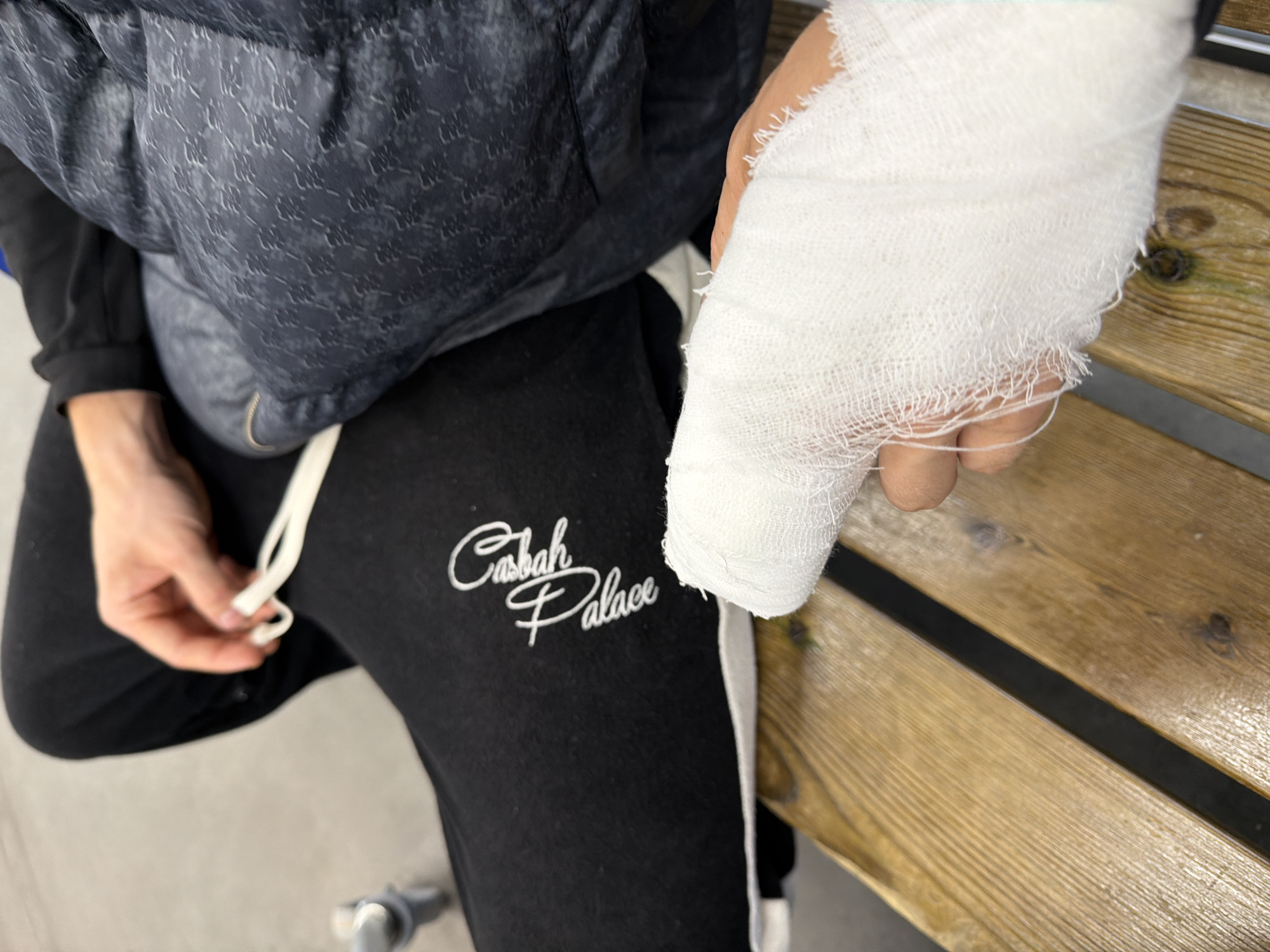 The image shows a close-up view of a person's bandaged hand resting on a wooden bench. The person is wearing black sweatpants with white lettering that appears to say "Casbah Palace" and a dark, patterned puffer jacket. Their other hand is visible in the upper portion of the image, holding a white string. The background is blurred but suggests an indoor or covered public space, possibly a waiting area, with a glimpse of a metal object with wheels. The wooden bench is made of planks with visible grain and knots. The overall lighting suggests an indoor setting. No specific location in Nizhny Novgorod, Russia, can be identified from the image.