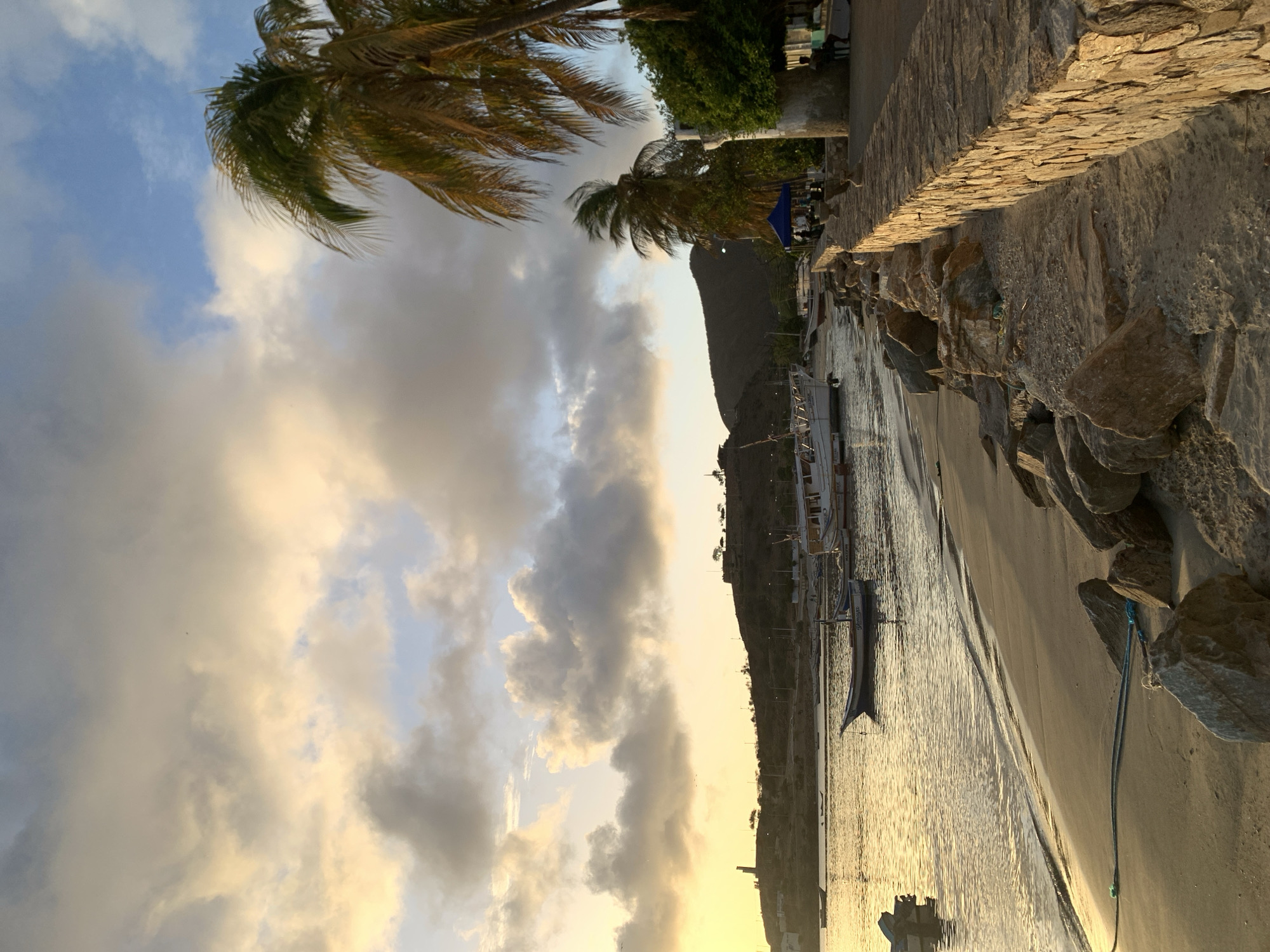 The image depicts a scenic view of the coastline in Juan Griego, Venezuela, during what appears to be either sunrise or sunset. The sky is filled with a dramatic display of clouds, tinged with hues of yellow and orange from the low-lying sun, contrasting with patches of blue sky. In the foreground, a sandy beach is dotted with large rocks. A thick, blue rope lies coiled on the sand near the water's edge.
The water is calm and reflects the warm light of the sky. Several boats are anchored in the bay; some are small, narrow vessels, while others are larger, more substantial boats with white hulls and visible masts.
Along the right side of the image, a stone wall runs parallel to the beach, forming a walkway or promenade. Palm trees with their fronds rustling in the breeze line the top of this wall. Further down the promenade, a small blue tent is set up, and a few people are visible, some sitting on benches under the shade of a tree.
In the background, a hilly landscape rises, with some structures and a prominent lighthouse-like building visible on one of the higher points. The overall impression is one of a tranquil coastal setting bathed in warm, atmospheric light.