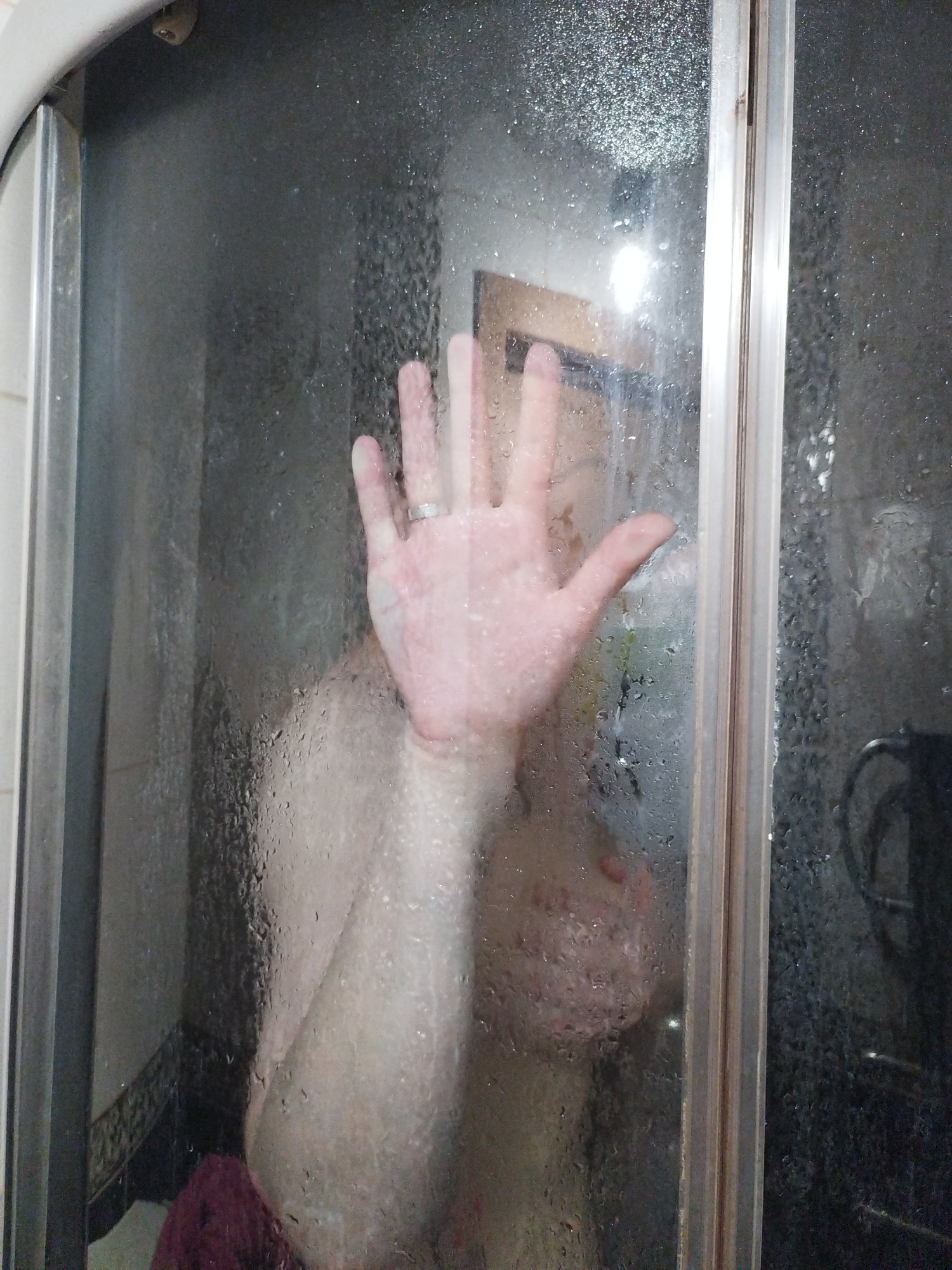 A person's hand with a ring is pressed against a steamy shower door. The person is partially visible behind the condensation, with their torso and chest visible. A dark red cloth is on the floor of the shower, and a tiled wall is visible in the background. The scene is inside a shower stall.