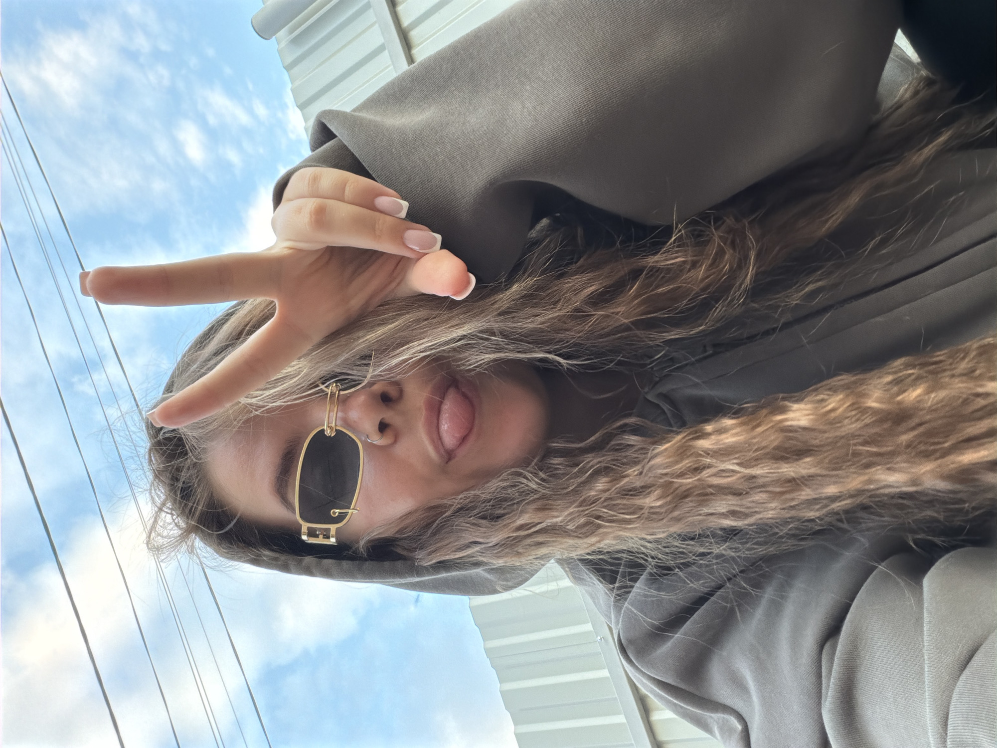 A young woman is positioned in a low-angle selfie with a view of the sky and utility wires visible above. The woman wears a dark gray hoodie with the hood up and large, black and gold sunglasses, she is sticking her tongue out and giving a peace sign with her right hand. Her long, wavy brown hair is loosely arranged around her face. The background includes a metallic structure above. There is no clear evidence to confirm the location is in Chaikovsky, Russia.
