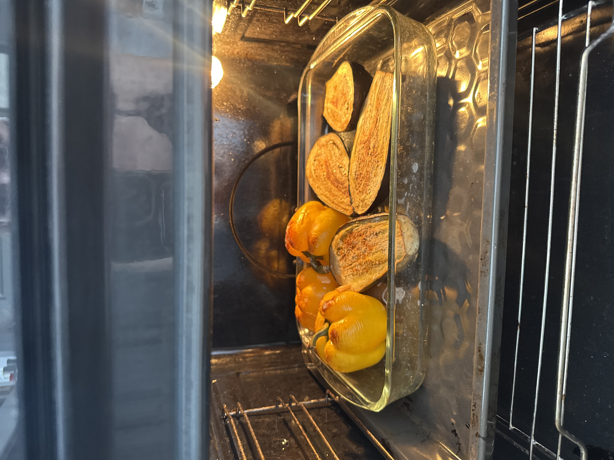 The image depicts the interior of an oven, located in Sochi, Russia, with food actively roasting inside. A clear rectangular glass baking dish rests on a metal oven rack, containing multiple vegetables. Three yellow bell peppers, largely intact but showing signs of blistering and light charring from heat, are visible. Adjacent to the peppers are four halves of eggplant, split lengthwise and positioned cut-side up, exhibiting browned and softened flesh from the roasting process. A bright incandescent light source, originating from the upper right interior of the oven, illuminates the scene. The oven's dark metallic interior walls show some grease and residue buildup. Additional metal oven racks are visible below the baking dish and along the left interior wall. The top edge of the oven's inner frame is partially visible at the top of the image. No individuals are present in the frame.