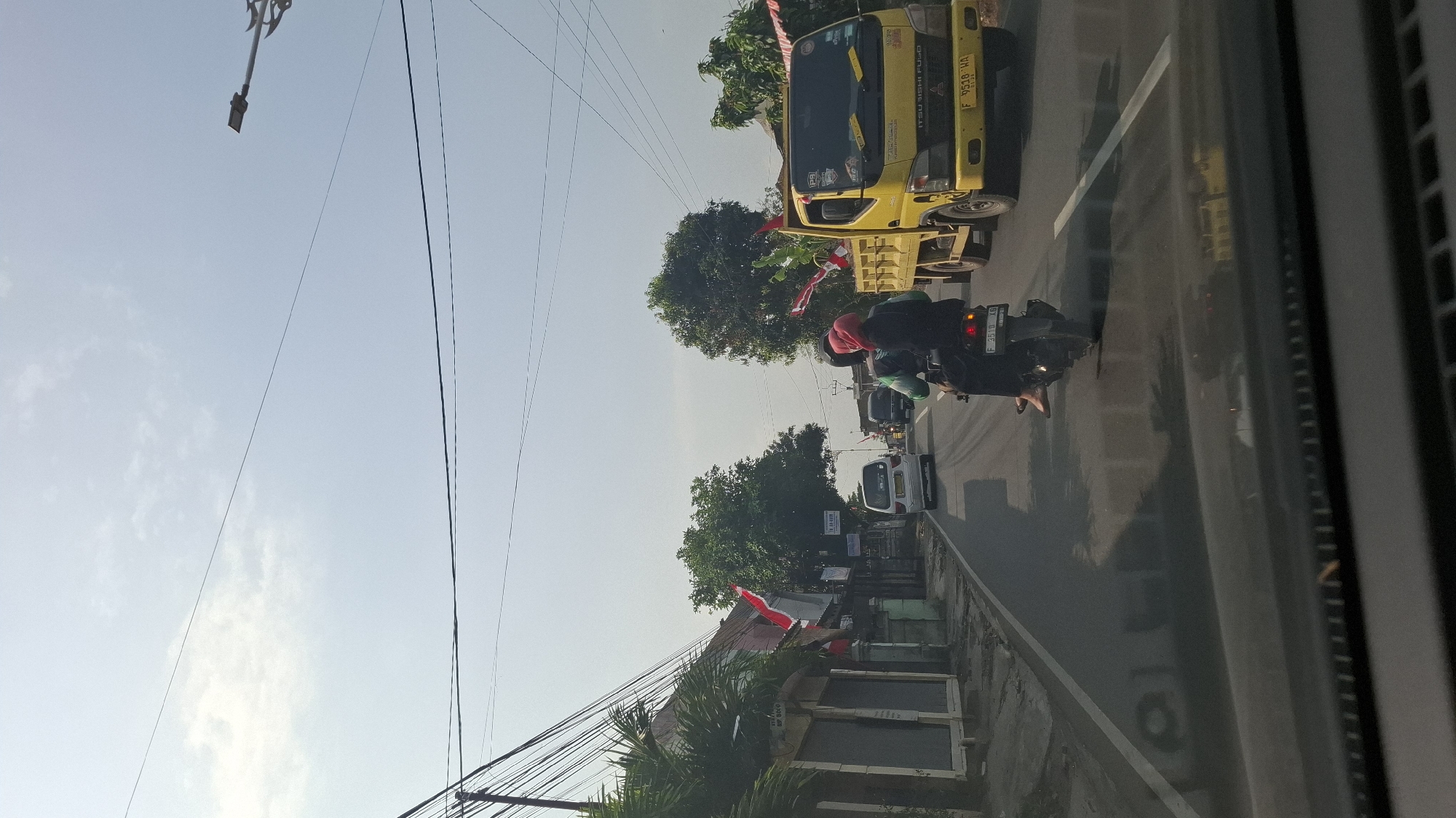 This is a vertical, eye-level, medium shot taken from inside a vehicle looking out onto a road in Cilaku, Indonesia. The scene depicts a street with moderate traffic under a clear, bright sky. On the left side of the road, there are several buildings with closed shutters, and palm trees are visible in the foreground. Red and white Indonesian flags are displayed along the side of the road and on the trees.
The road itself is asphalt and has lane markings. Several vehicles are present: a white minivan, a black motorcycle with two passengers (one wearing a red headscarf), and a yellow truck. The motorcycle is in the foreground, with the passengers facing away from the camera. The yellow truck is to the right of the motorcycle, and the white minivan is further down the road on the left. There are power lines crisscrossing the sky above the scene, and a street light is visible on the right side of the frame. The overall impression is of a typical day on a busy road in an Indonesian town.