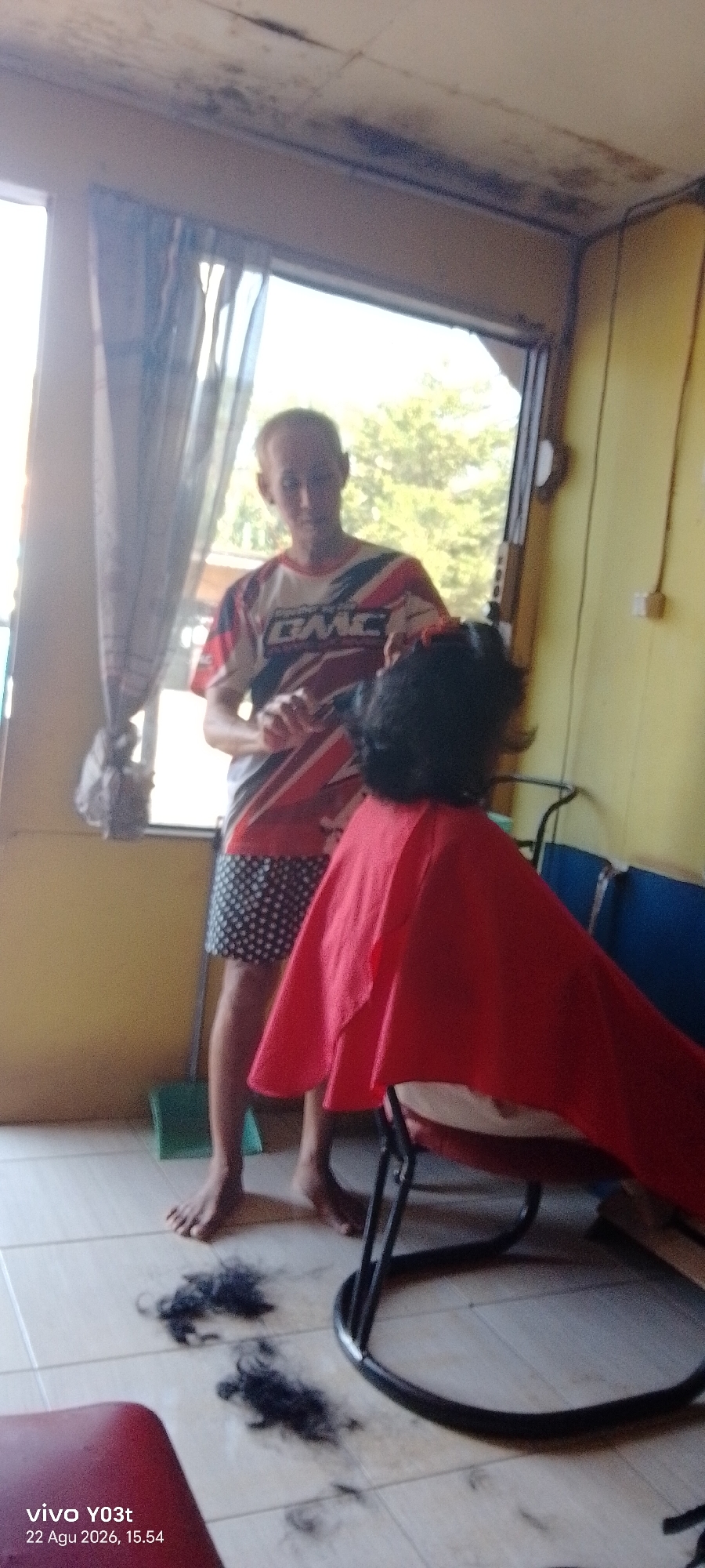 This is a medium shot of a man in a t-shirt and shorts giving a haircut to a person wearing a red cape in what appears to be a simple barbershop in Dawidawi, Indonesia. The man, who has light skin and short hair, is standing and appears to be using scissors and a comb. The person receiving the haircut is seated in a chair covered by a red cape and their back is to the camera. Clippings of dark hair are scattered on the tiled floor in the foreground, indicating the haircut is in progress. The background shows a window with curtains, a yellow wall, and some blue chairs. The overall setting suggests a casual, possibly outdoor or semi-outdoor, establishment.