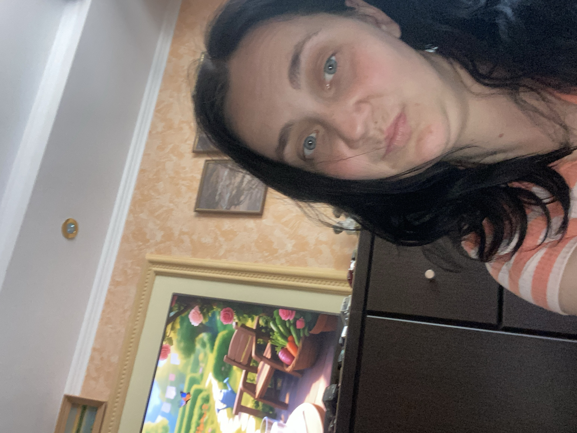 A woman with dark hair and blue eyes is pictured in a selfie from the chest up. She is wearing an orange and white striped shirt. Behind her, on the wall, is a framed painting of a garden scene. The painting depicts a wooden chair, a basket of vegetables, and flowers. The wall has an orange, textured wallpaper. A dark brown cabinet is visible below the painting. The lighting appears to be indoor lighting, with a ceiling light fixture visible in the upper part of the image. The location is unknown.