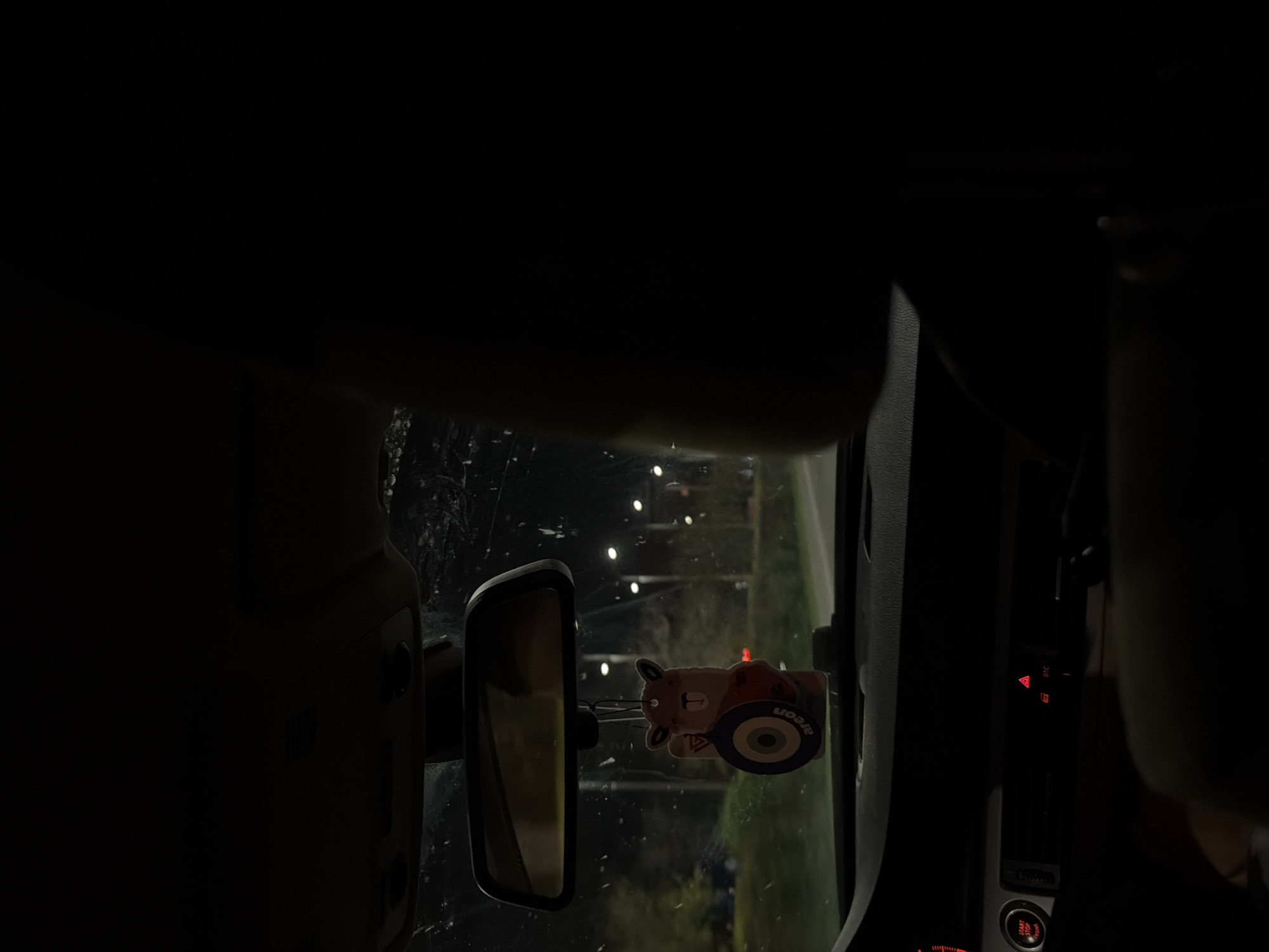 The image captures the interior of a car at night, viewed from what appears to be the driver's perspective, looking through the windshield. The scene is predominantly dark, contrasting with the bright artificial lights outside the vehicle.
A prominent feature in the center-left is the car's rearview mirror. Hanging from the mirror is a small, cartoon-like air freshener, resembling a brown animal figure in an orange outfit sitting on a blue wheel. The brand name "areon" is clearly visible on the air freshener.
Through the car's windshield, which exhibits some visible streaks or smudges, several bright, circular lights are arranged in a vertical pattern in the background. These are likely streetlights or other overhead illuminations in an urban or suburban environment, such as Plovdiv, Bulgaria.
On the right side of the frame, parts of the car's console or dashboard are visible, illuminated by red backlighting. A red triangular hazard warning light is clearly active, and below it, the letters "DTC" are partially discernible. Further down, a "START STOP ENGINE" button is also illuminated in red. The overall impression is a quiet, nocturnal scene from inside a parked or slowly moving car. No people are visible in the image.