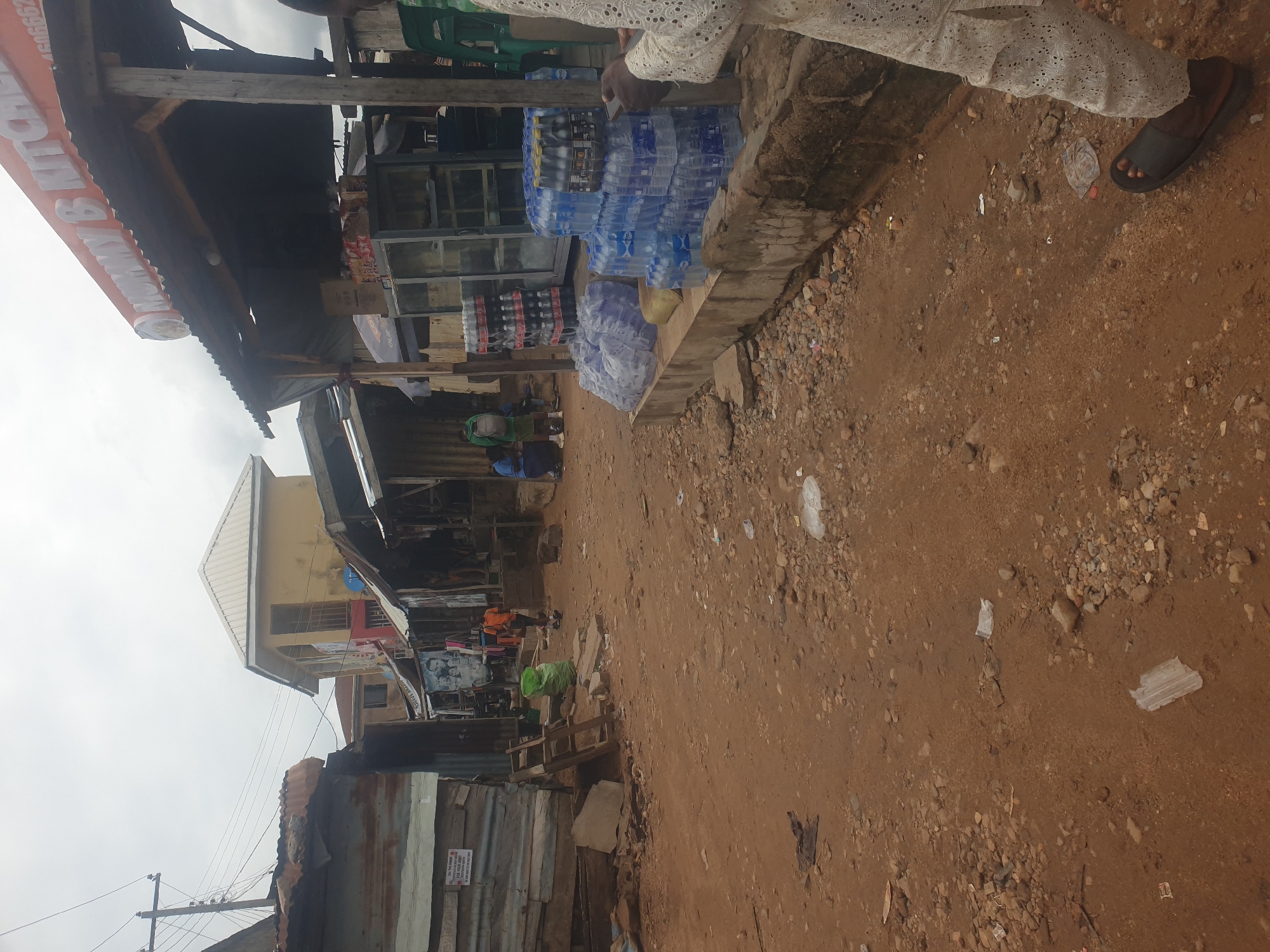 The image shows a street scene in an unidentified city. Along one side are shops, including one selling water bottles and another with a sign reading "B. KITCHEN & B." The street is unpaved and littered with trash. A person wearing white clothing and sandals is standing near the water bottle shop. In the background, buildings of varying heights can be seen. The sky is overcast.