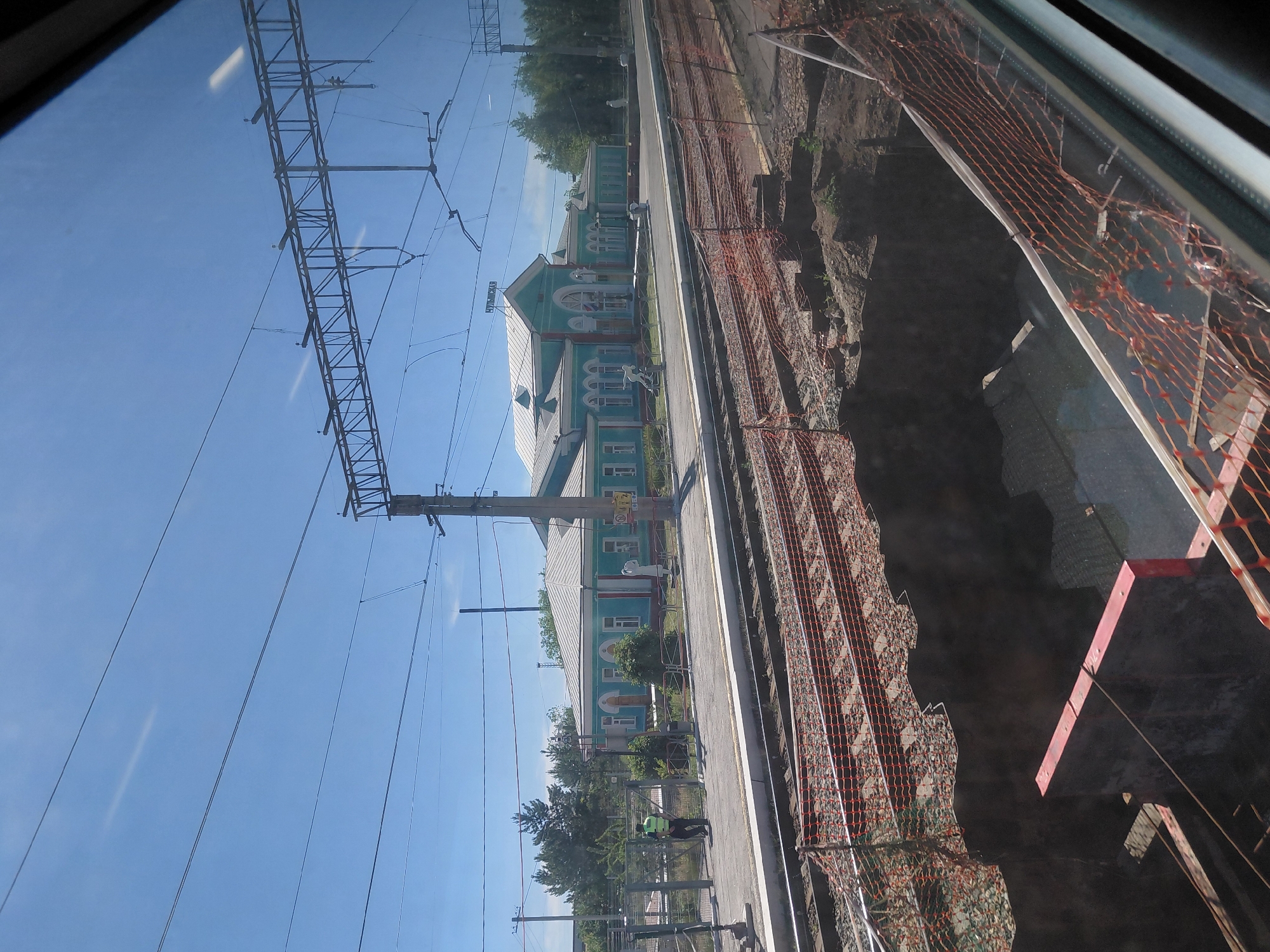 This is a medium-shot, slightly angled exterior view of the Chulym railway station in Chulym, Russia, taken on a clear day. The foreground shows construction work with orange netting fencing off a deep trench next to the railway tracks. The trench appears to be a work in progress, with exposed earth and some construction materials visible.
The middle ground features a light blue, single-story building with a white roof, identified as the railway station by a sign reading "ЧУЛЫМСКАЯ" (Chulymskaya) in Cyrillic lettering at the top of the main entrance. The building has arched windows and doors, and two white statues are placed outside on either side of the entrance. A paved platform runs in front of the station, and a person in a high-visibility vest is seen walking on the platform near a fence.
The background consists of trees and utility poles supporting overhead railway power lines. The sky is bright blue with a few faint white streaks, possibly reflections on the window through which the photo was taken. The overall scene depicts an active railway station undergoing some form of renovation or construction.