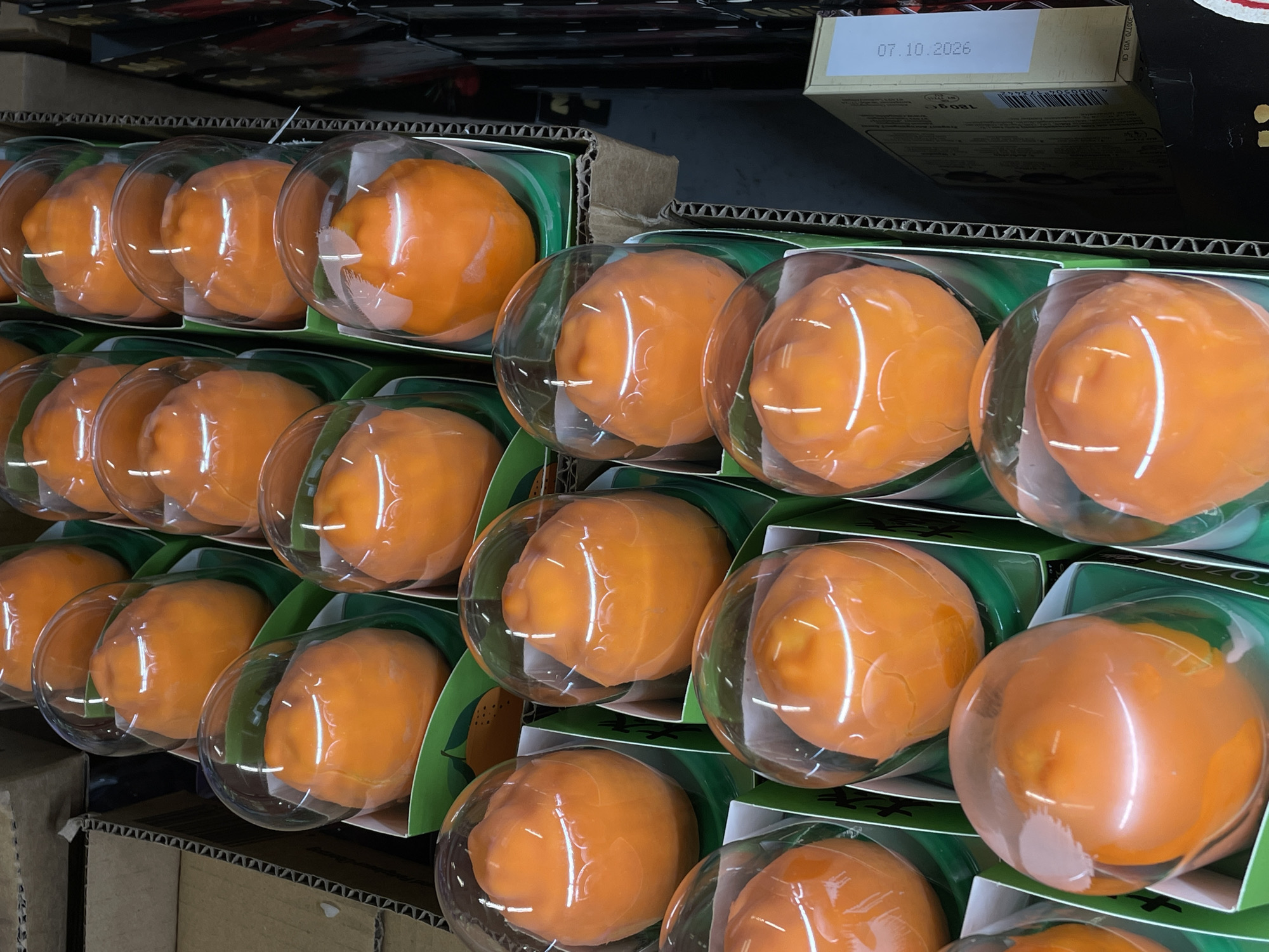 This is a close-up, eye-level, interior shot of a retail display case containing multiple, individually packaged, bright orange persimmons in Siegburg, Germany. The persimmons are round and smooth, with a slight indentation at the top, and are encased in clear plastic domes. Each persimmon sits in a green, cardboard holder with a stylized orange graphic visible on some. The display is arranged in rows and columns within a corrugated cardboard box. The lighting is artificial and even, highlighting the glossy surface of the persimmons and the plastic packaging. In the background, other boxes and products are visible, along with the texture of the cardboard display. A portion of a product label with the text "07.10.2026" is visible on the right side of the image.