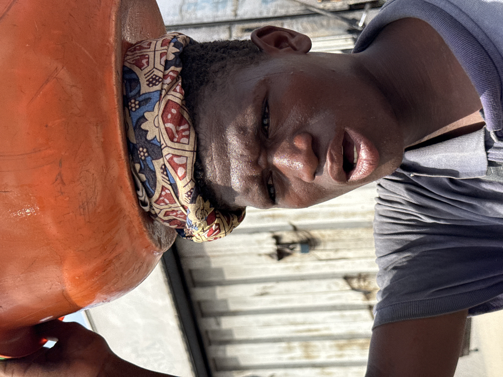 A person is pouring liquid from an orange bucket onto their head. The person has dark skin and is wearing a printed headscarf and a grey shirt. The background appears to be a corrugated metal surface. This likely takes place in Lagos, Nigeria.