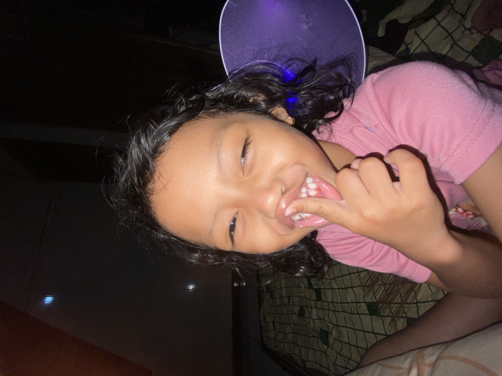 The media file features a close-up, tilted shot of a young child, likely a girl, with dark, curly hair. She is wearing a pink t-shirt. The child is lying down, with her head tilted back and her mouth open, showing her teeth. Her right hand is near her mouth, with her fingers touching her lips. Her eyes are open and she appears to be smiling or laughing. A portion of a purple object, possibly a hat or a toy, is visible behind her head, with a blue light emanating from it. The background is dark, suggesting an indoor setting at night. A patterned blanket or bedspread is visible beneath the child. The overall mood is playful and happy. No visible text is present in the image. The location context is Gelam, Indonesia.