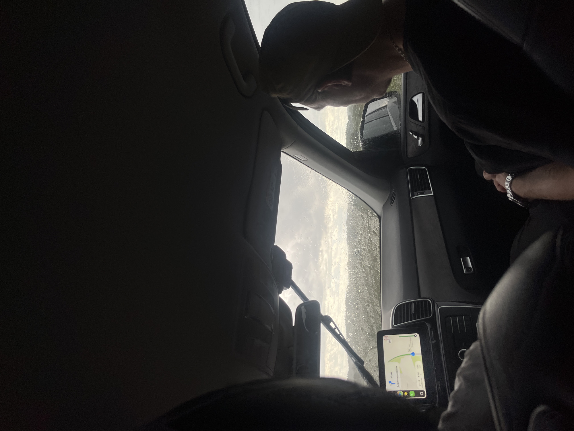 The image captures the interior of a car, looking forward through the windshield. Rain is visible on the glass, distorting the view of a landscape of hills and trees under a cloudy sky. A man wearing a black t-shirt and a baseball cap is seated in the passenger seat, looking out the window. On the dashboard, a navigation screen displays a map with route information. The overall lighting suggests an overcast or rainy day. There is no specific information in the image to confirm or deny the location as Krasnoyarka, Russia.