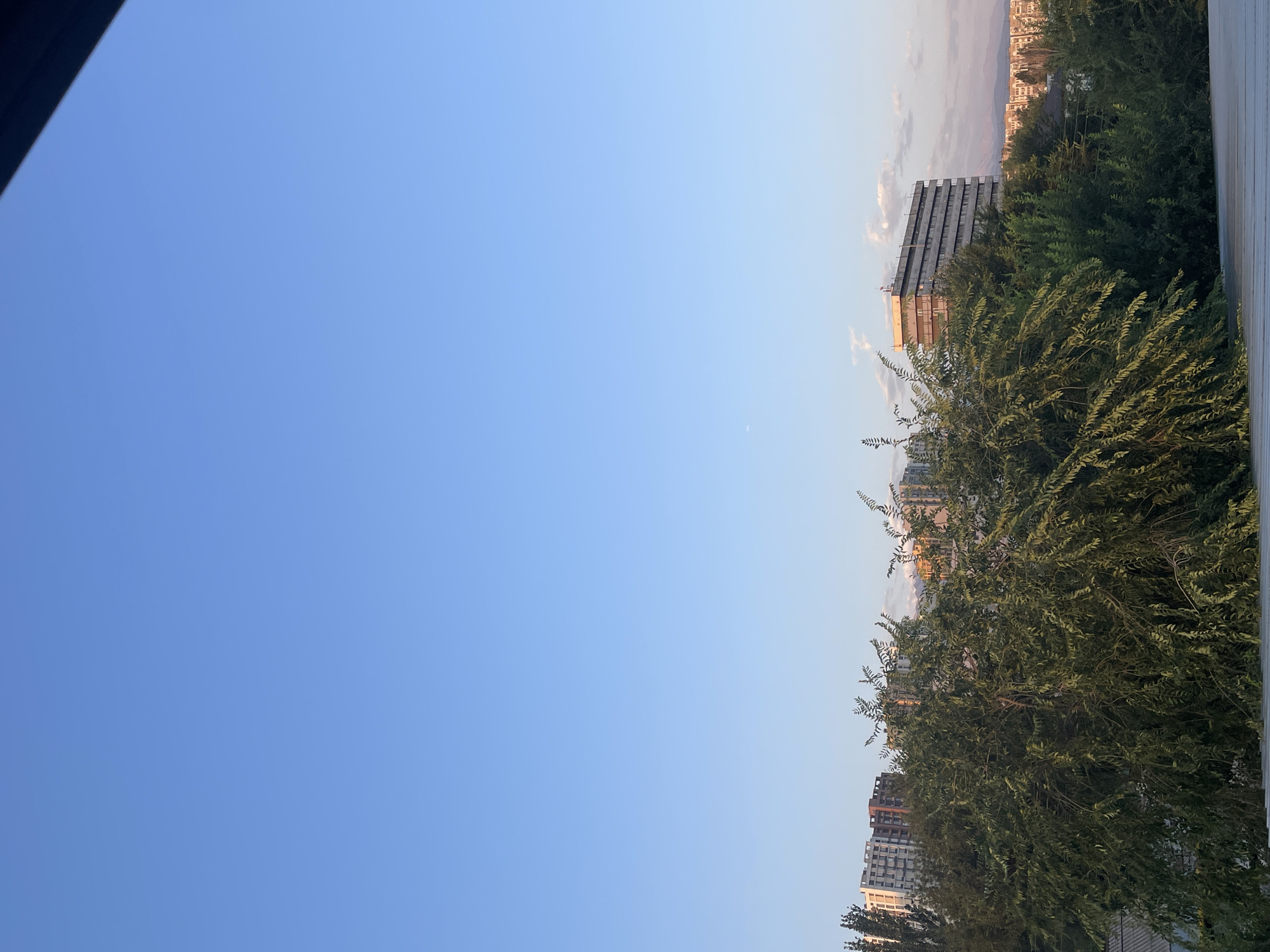 The image shows a bright, clear sky above a cityscape, with a prominent building and trees in the foreground. The sky is a soft, pale blue, with a faint crescent moon visible. Below the sky, the tops of several apartment buildings are visible, with a large, rectangular building dominating the right side of the frame. The buildings appear to be made of concrete and glass, with many windows. Lush green trees with dense foliage fill the lower half of the image, obscuring some of the lower parts of the buildings and a residential area with a gray roof. In the distance, mountains are faintly visible, possibly the Tian Shan mountains that surround Almaty. The lighting suggests it is either early morning or late afternoon, with a warm glow on some of the buildings. The scene depicts a typical urban environment in Almaty, Kazakhstan.