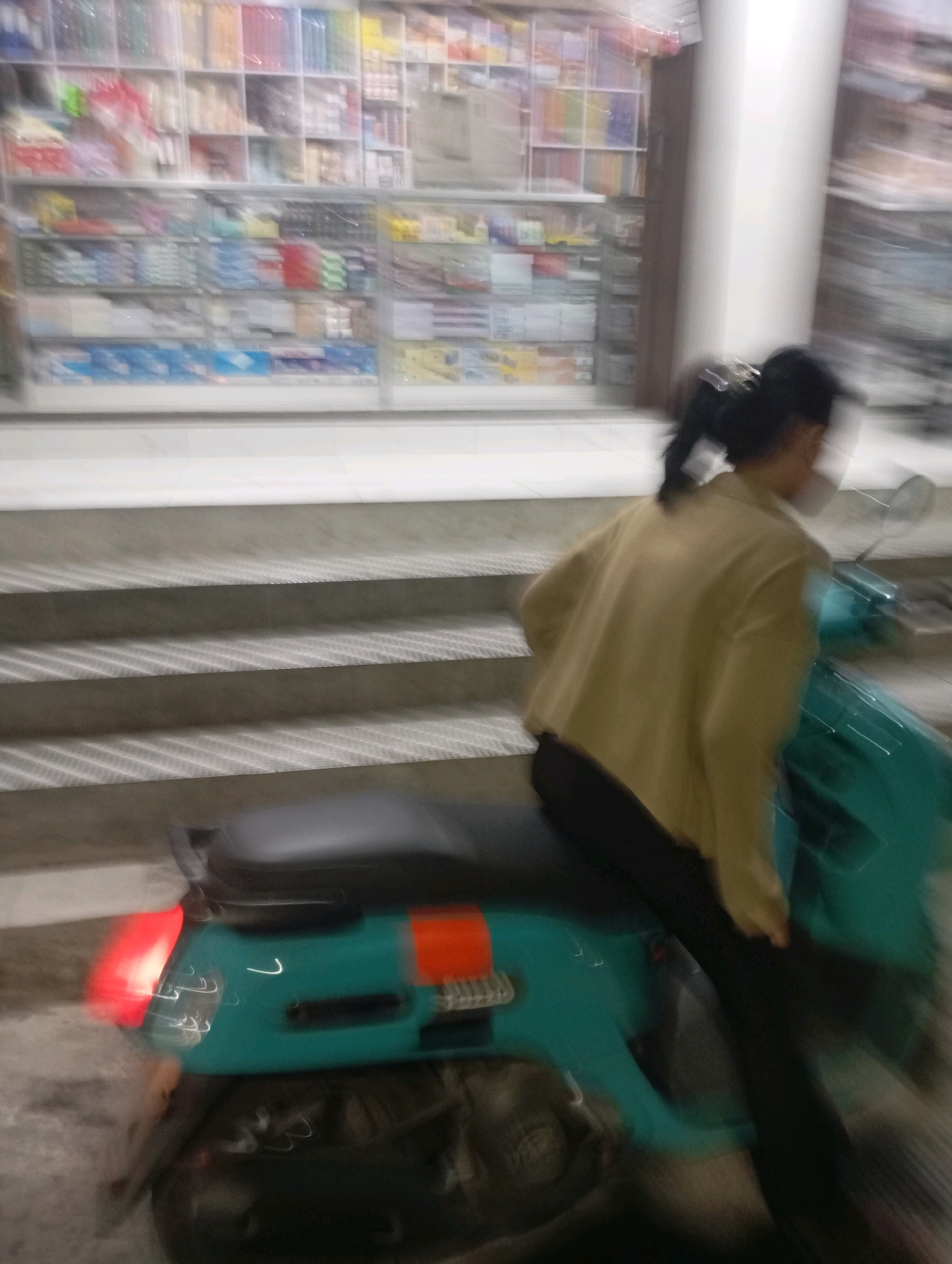 In Babelan, Indonesia, a person is on a turquoise scooter. The person is wearing a light brown jacket and dark pants. The scooter is in motion, evidenced by motion blur in the image, and is positioned on a set of stairs. The background shows shelves filled with various items, possibly indicating a store or market. The lighting suggests it is nighttime or dimly lit.