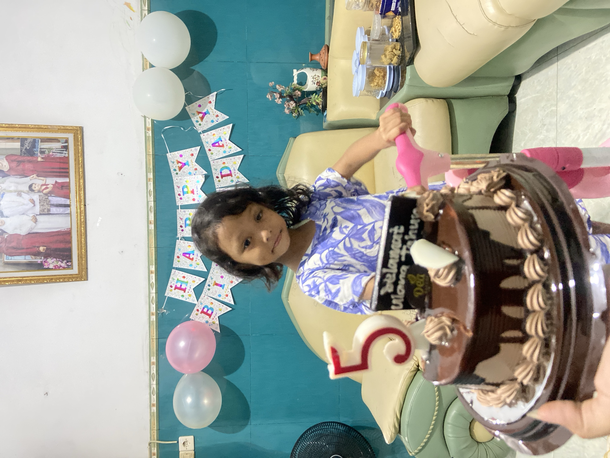The scene captures a birthday celebration in Gelam, Indonesia. A young girl wearing a blue and white patterned shirt is positioned in front of a chocolate cake. The cake is decorated with a "5" candle, a sign that reads "Selamat Ulang Tahun," and small chocolate cream designs. The girl holds a pink object, possibly a toy hammer.
In the background, "HAPPY BIRTHDAY" banners are hung on a blue-green wall, flanked by balloons, two white and two pink. Behind the girl, there is a tan and green colored upholstered armchair. On the upper right corner there are jars of snacks on top of the arm chair. On the left side, a framed picture hangs on the wall. A standing fan is visible in the bottom center of the frame. The environment suggests an indoor party at a home.