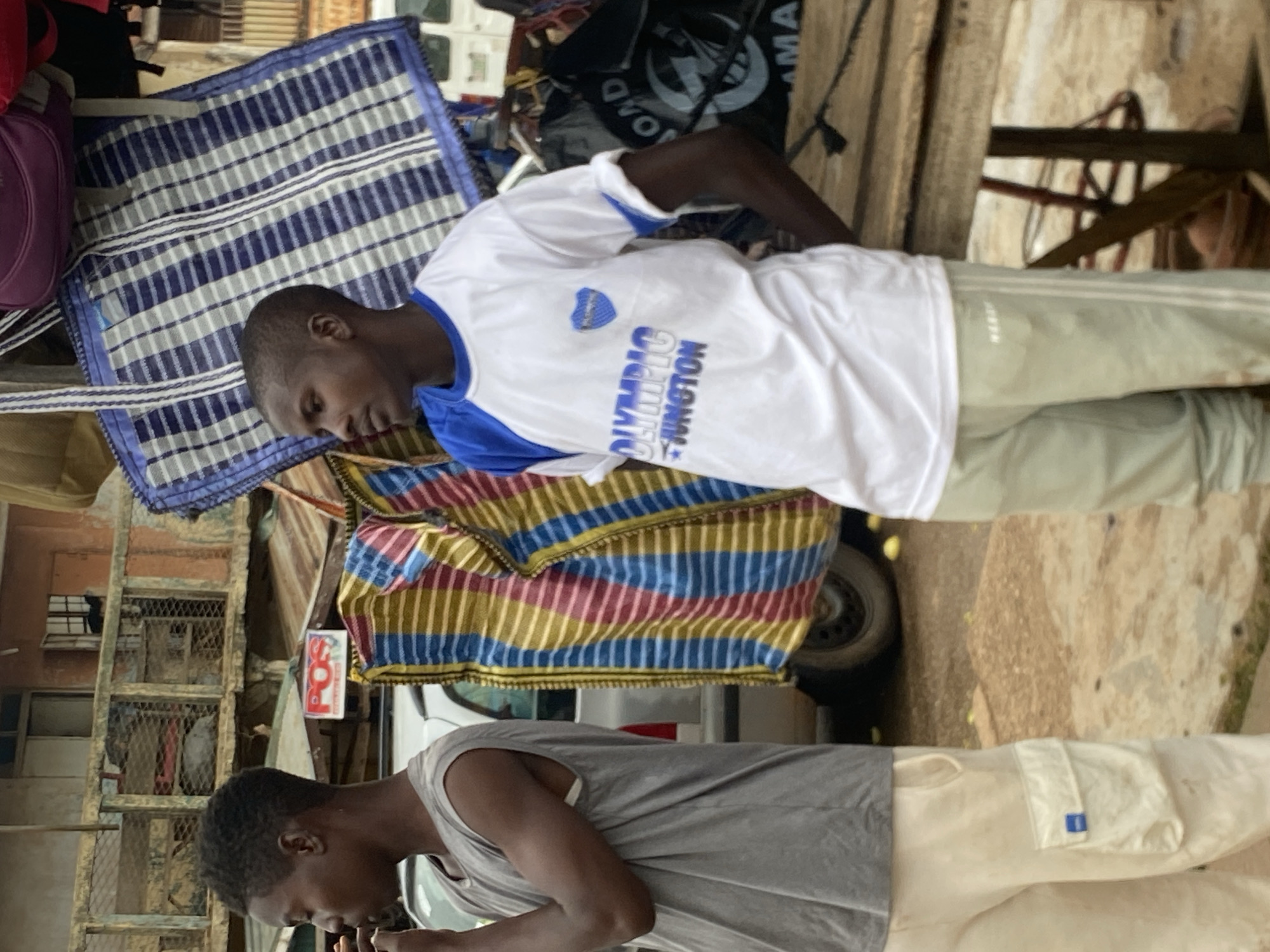 In Bukuru, Nigeria, a young Black male stands in profile facing left, his right hand raised towards his mouth. He wears a sleeveless gray t-shirt and light-colored cargo pants with a blue logo on the right pocket. To his right, another young Black male faces forward and slightly to the left, wearing a white t-shirt with blue accents and blue lettering that reads "OLYMPIC JUNCTON" with a blue shield logo. He also wears light-colored pants with white stripes down the sides. Behind the two individuals are various market stalls and structures, including a vehicle, colorful striped fabrics hanging, and bags. A sign with "POS" in red letters is visible above a stall to the left. The ground is a mix of dirt and possibly paved areas.