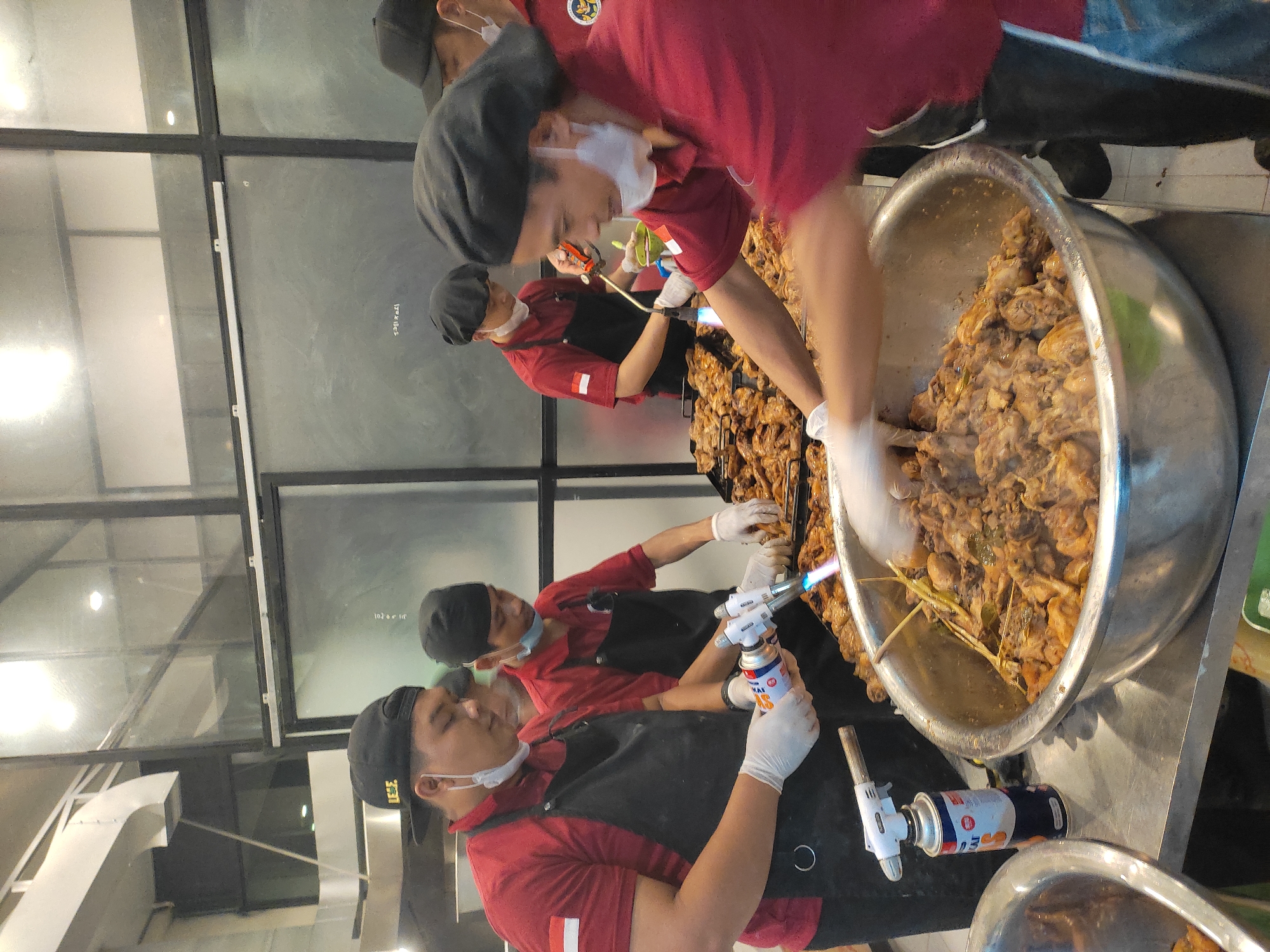 The image depicts a group of men, likely chefs, preparing food. The scene appears to be an indoor food stall or a kitchen area, possibly in a market or restaurant. One man in the foreground, wearing a red shirt and black apron, is using a blowtorch on a large metal bowl filled with cooked food, possibly chicken. He is also wearing white gloves and a face mask. To his left, another man in a similar outfit is also holding a blowtorch. Behind them, other men in red shirts and black head coverings are visible, also engaged in food preparation. The environment includes stainless steel surfaces, grills with cooked food, and large metal bowls. The presence of Indonesian flags on some of the uniforms suggests the location is Indonesia. While the image does not explicitly show elements of the city of Ponorogo, Indonesia, the context of food preparation and Indonesian attire hints at a possible location within the country.