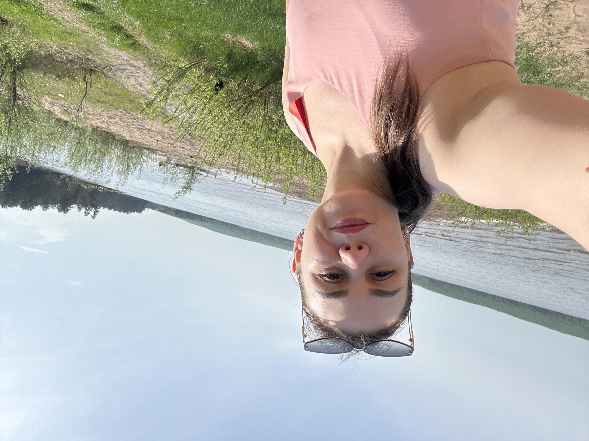 A woman with sunglasses perched on her head is facing the camera. She is wearing a light pink top. The background shows a river and a sandy shore with some greenery. The picture appears to be taken in a sunny, outdoor setting. The image appears to have been taken at a location near the water in Chaikovsky, Russia.