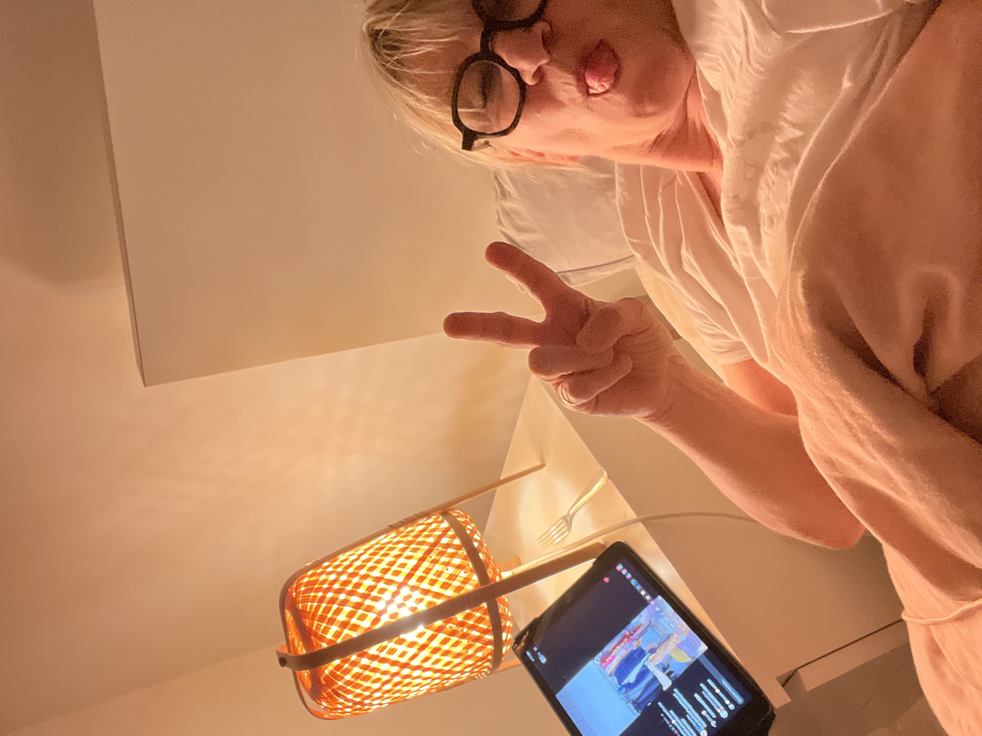A woman with short blonde hair, wearing glasses and a white shirt, is sticking her tongue out and making a peace sign. She appears to be lying down, judging by the pillow behind her head. A woven lamp and a tablet are positioned below her, along with a fork. The location is Chambœuf, France.