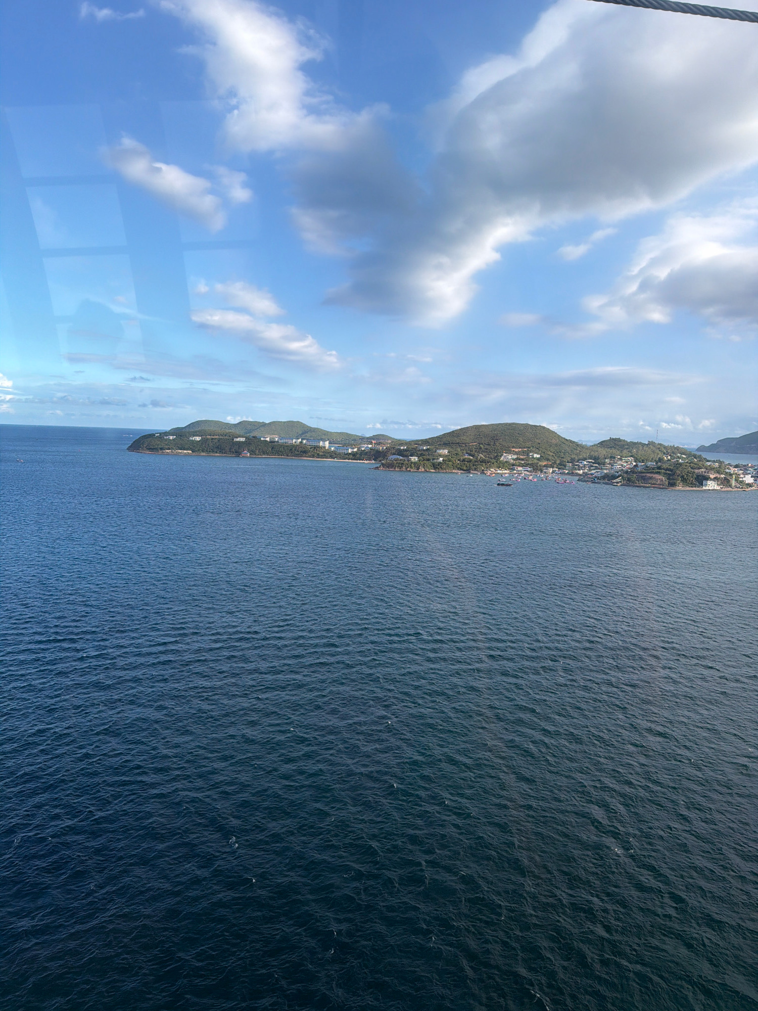 This image depicts a broad, blue ocean with gentle waves, stretching towards a distant coastline. The coastline is characterized by green, rolling hills dotted with white buildings and a marina filled with numerous boats. The sky above is a vibrant blue, featuring scattered white and gray clouds. The overall scene suggests a tropical or coastal location with developed areas along the shore. The location is not Nha Trang, Vietnam.