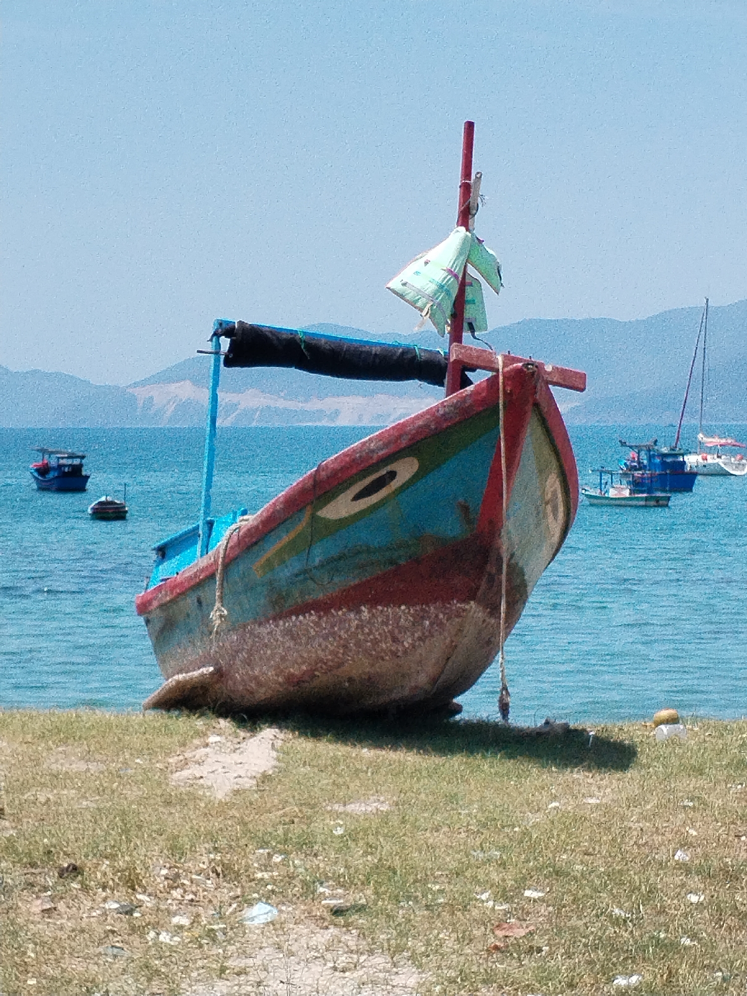 A beached, weathered fishing boat dominates the foreground in what appears to be Bac Nha Trang Ward, Vietnam. The boat's hull is a mix of red, blue, and tan colors, with a single mast displaying a scrap of light green material. The boat rests on sandy and grassy terrain littered with debris. In the background, the turquoise water of the bay contains several other boats of varying sizes, including a sailboat. Hazy mountains are visible on the horizon under a clear, light blue sky.