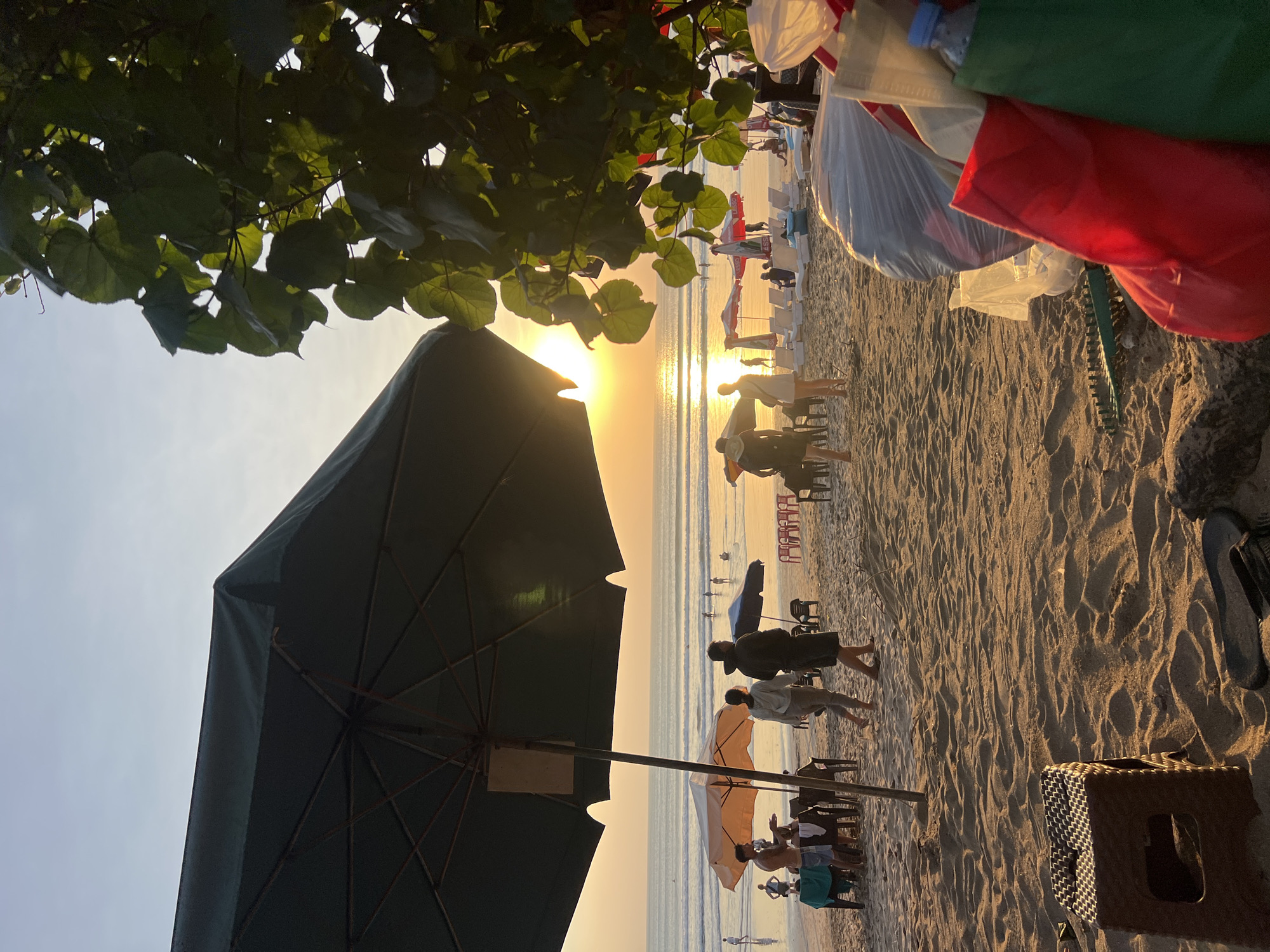 The image depicts a beach scene in Kuta, Indonesia during sunset. The sun is low on the horizon, casting a warm golden glow across the ocean and sand. Several people are visible on the beach, some walking, some sitting under umbrellas, and others in the water. There are also beach chairs and tables set up, suggesting a commercial area. In the foreground, a large green umbrella partially obscures the view, and to the right, a cluster of colorful shopping bags and trash are piled up. Lush green foliage from a tree frames the right side of the image. The sand is marked with footprints and the gentle waves of the ocean are visible in the background.