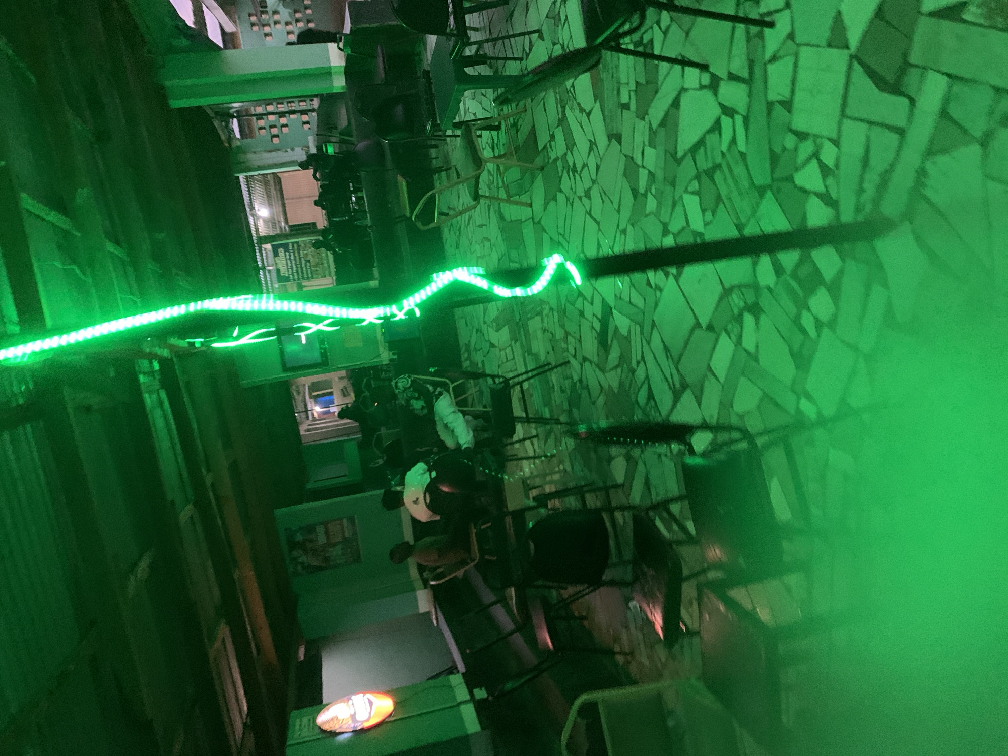 The scene depicts an interior or semi-outdoor social establishment in New Stadium, Nigeria, dominated by intense green artificial lighting. A bright string of green LED lights forms a prominent, curving vertical line in the foreground and mid-ground, extending from the upper to the lower frame. The floor consists of a mosaic pattern of irregularly shaped, light-colored tiles or concrete fragments.
Approximately 6-8 adult individuals are visible, primarily seated at small, dark tables equipped with simple chairs, in the mid-ground and background. One person in a white t-shirt and another in a dark shirt are clearly discernible in the mid-left. Their postures suggest casual gathering or conversation.
Along the left wall, an oval-shaped, orange and white illuminated sign, possibly a brand logo, emits a glow, though its colors are heavily influenced by the pervasive green light. Further along this wall, a framed poster or sign is affixed, and a dark, elongated surface, possibly a counter or bench, runs horizontally. A television screen with a blue tint is partially visible on the far left. The ceiling exhibits exposed wooden beams and planks, indicating an unadorned or industrial aesthetic. In the background, an opening reveals an outdoor area or adjacent space with faint ambient light.