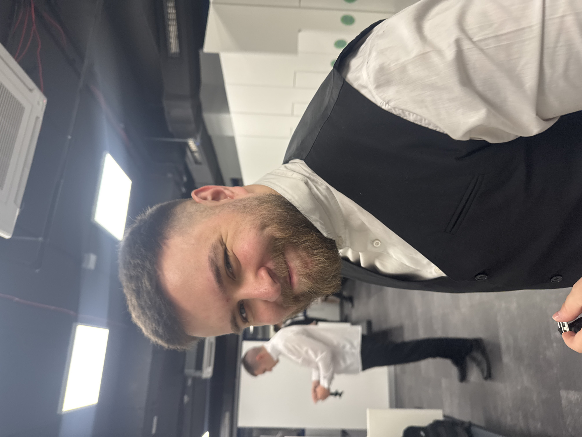 A man with a beard and short hair wearing a white shirt and black vest is standing in a room with a black ceiling and white walls. He is smiling and looking at the camera. Another man in a white shirt and black pants is visible in the background. The photo appears to be taken indoors, possibly in a commercial or office space. A small, black object is in the man's hand. There is no evidence to connect this image to Sofia, Bulgaria.