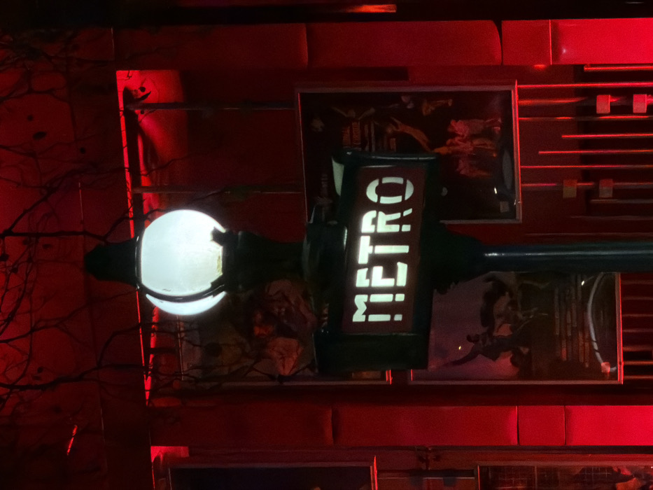 A brightly lit 'METRO' sign sits beside a globe lamp, centered in a red-toned setting. The sign's letters are bold and illuminated. The background consists of rectangular patterns and framed artworks. The overall scene has a staged or artificial aesthetic, with a focus on the sign as a central element within the elaborate display.