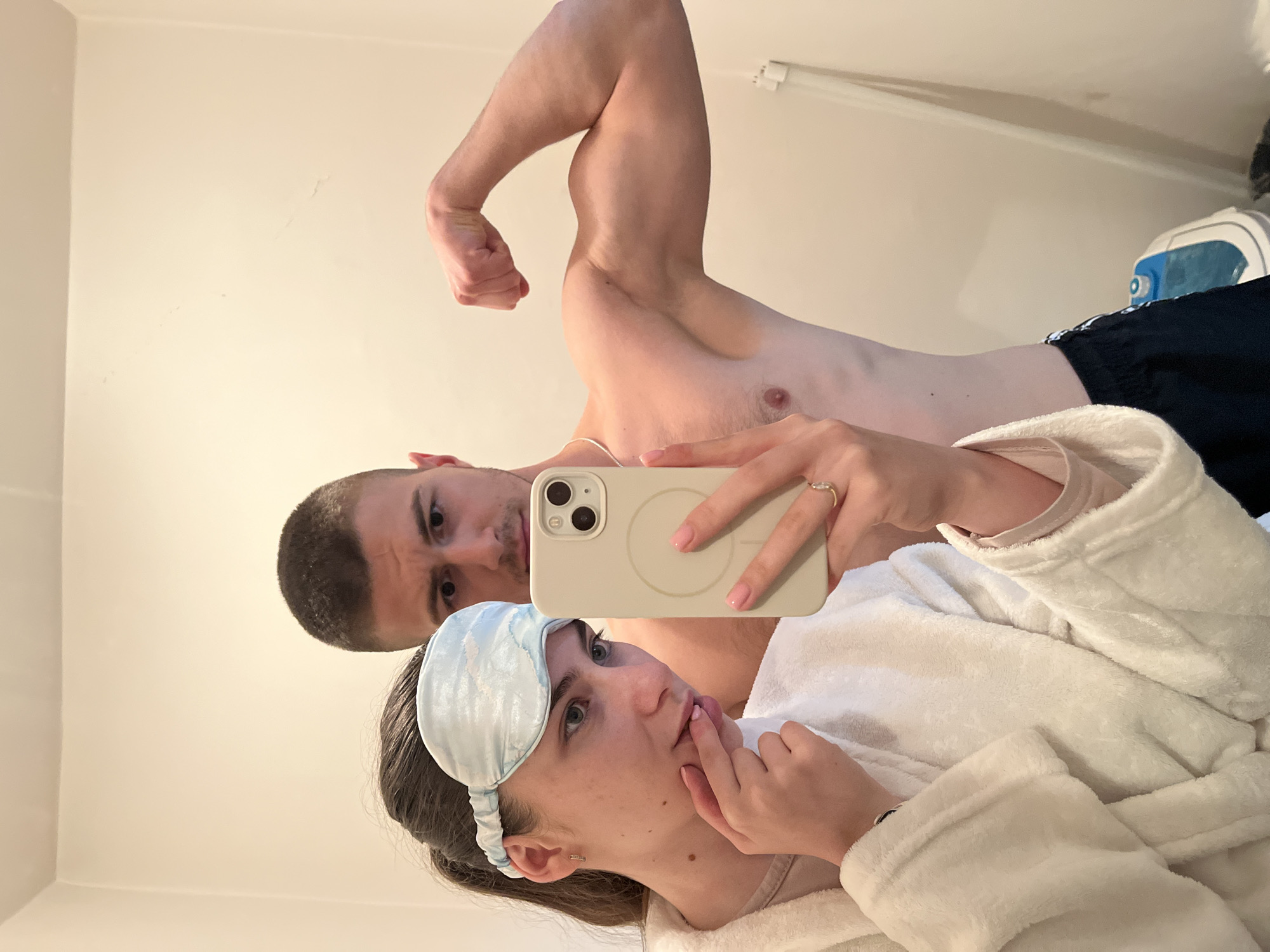 A photograph taken indoors depicts two individuals, a male and a female, in an Unknown, Unknown location. The female is wearing a white bathrobe and a blue eye mask and has her finger to her lips. The male is shirtless, flexing his bicep, and holding the phone. They appear to be taking a selfie, and the angle suggests the image was taken in a mirror. The background is a light-colored wall. An iron or steamer is visible in the top right corner of the image.