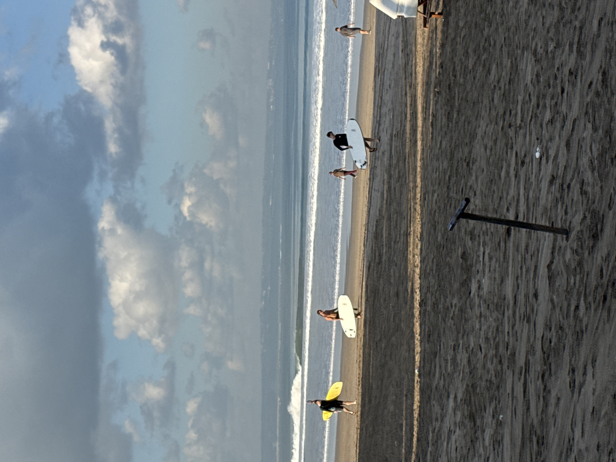 The image captures a daytime scene on Kuta Beach, Indonesia, featuring five individuals on a sandy shore with the ocean and a partly cloudy sky forming the background.
Three individuals are carrying surfboards and walking from left to right across the mid-ground. On the far left, a person dressed in dark attire carries a bright yellow surfboard. To their right, a shirtless individual in dark shorts carries a white surfboard under their right arm. Further right, a person in a black long-sleeved top and dark shorts carries another white surfboard under their right arm. Two additional individuals are visible closer to the water's edge: one in lighter-colored clothing walking away from the camera towards the right, and another in a dark t-shirt and shorts, also walking away from the camera on the far right.
The sandy beach, displaying ripples and footprints, extends from the immediate foreground to the water. The ocean features multiple small breaking waves closer to the shore, with calmer water stretching to a distant, hazy horizon. The sky is largely covered by large, grey and white cumulus clouds, with visible patches of light blue sky. In the foreground, a dark, T-shaped metal pole is embedded vertically in the sand towards the bottom-left. A portion of a white, possibly fiberglass, structure with a visible wooden base is present on the far right of the foreground.