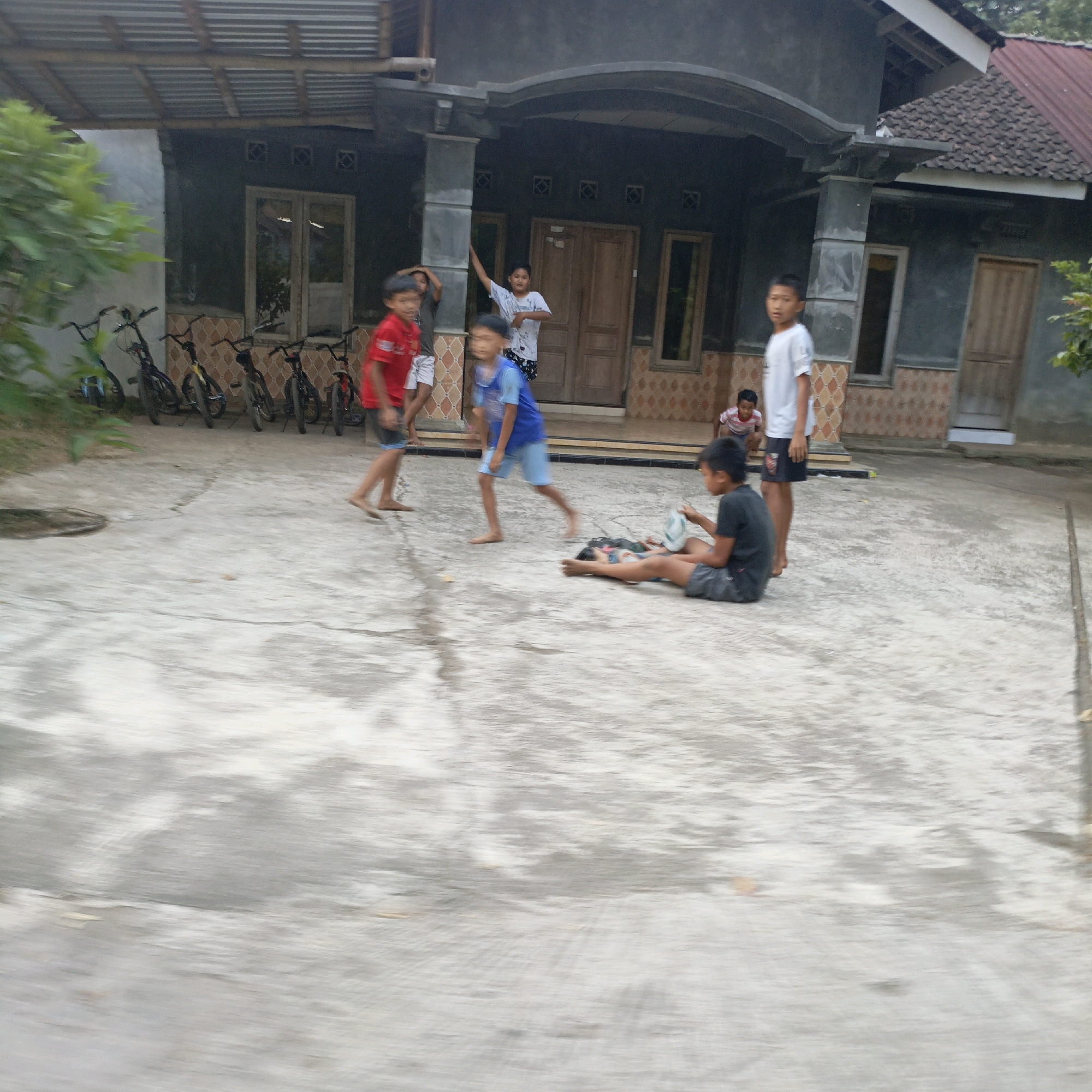In front of a house in Salatiga, Indonesia, a group of boys are playing on a concrete surface. The house appears to be a single-story structure with a dark grey facade and a tiled roof. Several bicycles are parked in a line near the house.
In the foreground, a boy in a dark t-shirt and shorts is sitting on the ground, holding a white object that appears to be a shoe. Another boy, lying on his back on a patterned mat, is near him.
To the left, a boy in a red t-shirt and shorts is in motion, his left leg raised as if kicking something. Next to him, another boy in a blue shirt and shorts is also in motion, with his arms outstretched. Further to the left, a boy in a red t-shirt and shorts is standing.
In the background, a boy in a white t-shirt is standing with his arms raised. Another boy in a white t-shirt and shorts is visible further back, near the house entrance. The overall impression is of a casual outdoor activity among children.