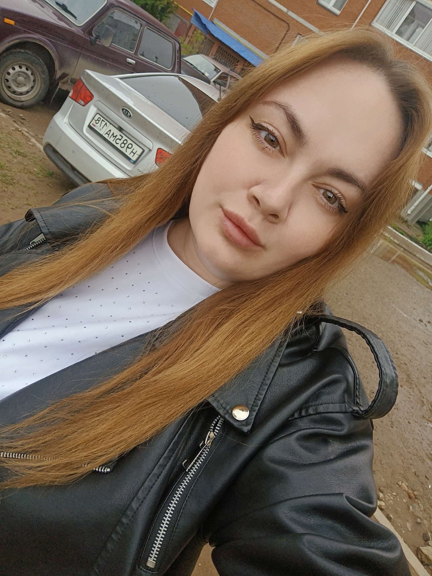 A young woman with long reddish-brown hair is taking a selfie outdoors. She is wearing a white t-shirt with small, sparkly embellishments and a black leather jacket, which is partially zipped up. In the background, there are two cars parked: a silver Kia and a burgundy SUV. Behind the cars, there's a multi-story brick building with windows. The ground appears to be wet, possibly after rain. The scene appears to be in the city of Chaikovsky, Russia, due to the license plate on the silver Kia.