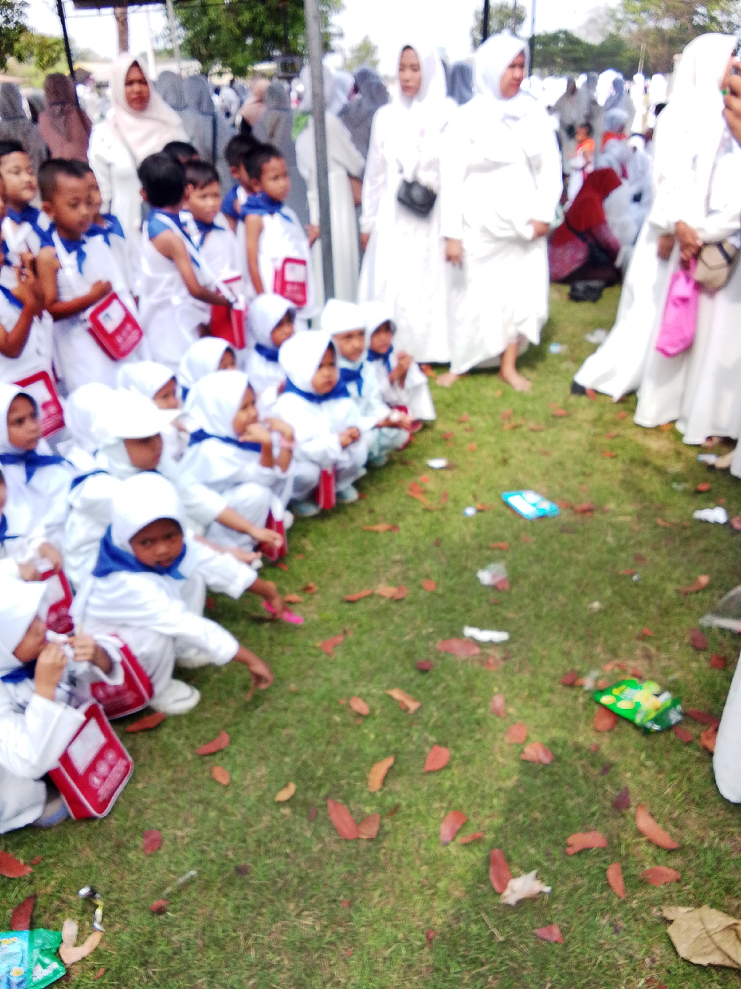 This is a high-angle, full shot of a group of children, mostly young boys, dressed in white uniforms with blue scarves and red pouches, sitting on a grassy field scattered with fallen leaves. Some adults, primarily women in white hijabs and dresses, are standing or sitting behind the children. The background is slightly blurred, showing more people and some trees. The scene appears to be an outdoor event, possibly a school gathering or religious ceremony. The presence of the "Masjid Raya At-Taqwa Cirebon" logo on some of the children's red pouches indicates the location as Cirebon, Indonesia, specifically in the vicinity of the At-Taqwa Grand Mosque. The overall impression is one of a community gathering, possibly related to religious education or a special occasion.