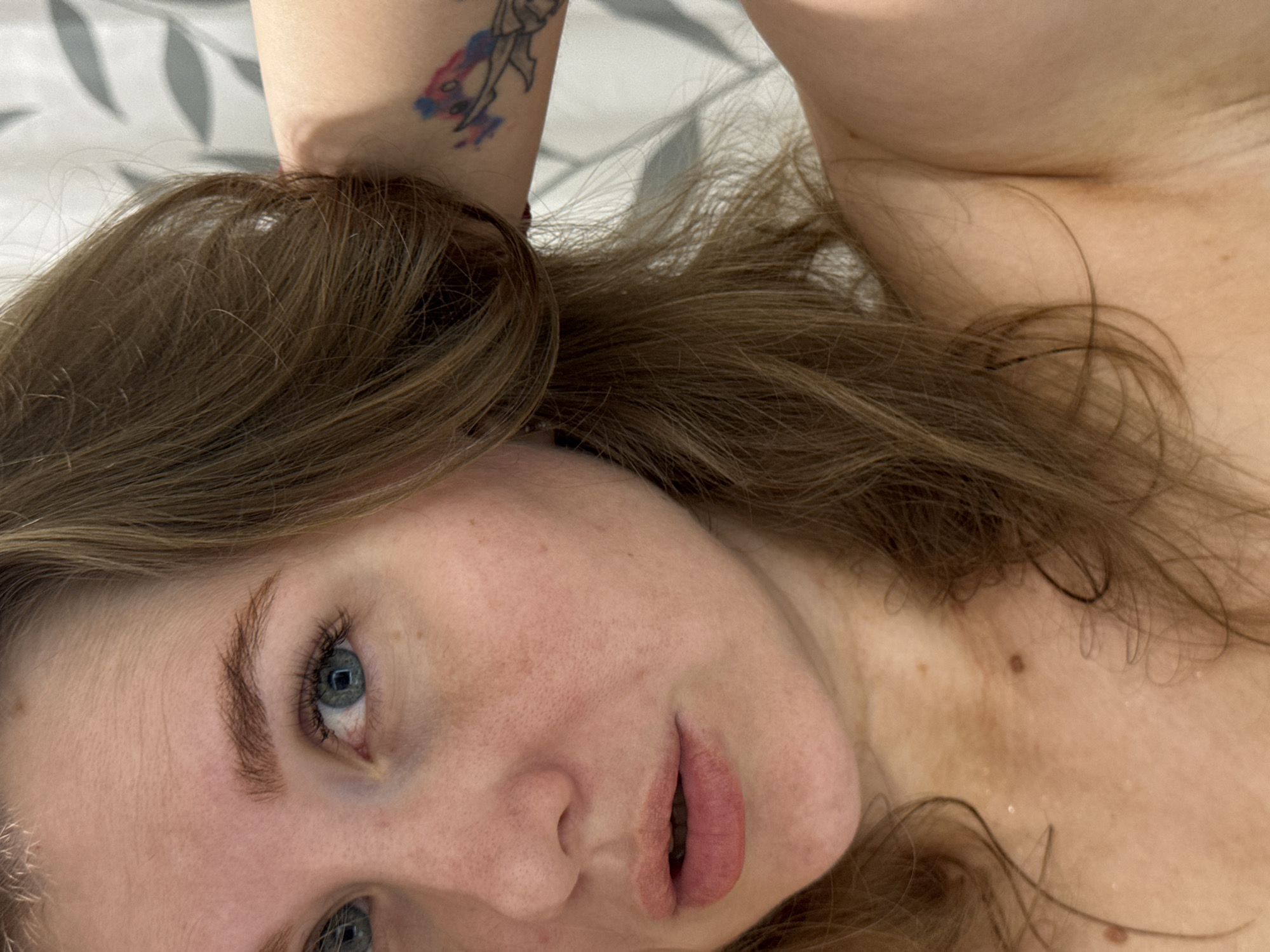 The image shows a close-up view of a woman lying down. She has light skin, blue eyes, and light brown hair. A tattoo is visible on her arm, which is raised above her head. The background appears to be a bed or a similar surface with a patterned sheet. There is no information available in the image to indicate its location in the city of Ферма, Russia.