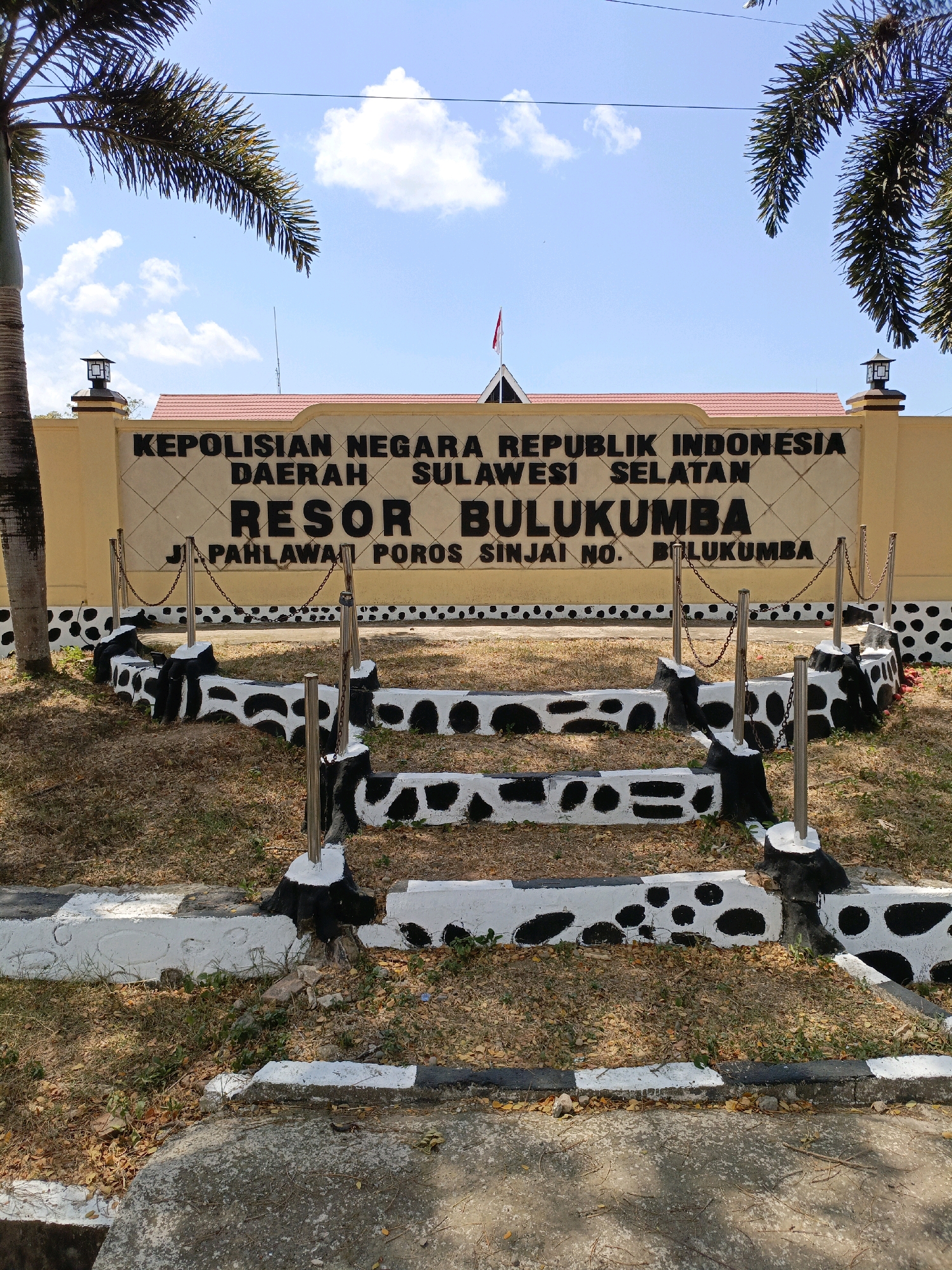 The image shows the entrance to the "KEPOLISIAN NEGARA REPUBLIK INDONESIA DAERAH SULAWESI SELATAN RESOR BULUKUMBA" in Bulukumba, Indonesia. The building is painted yellow with a red-tiled roof. A red and white Indonesian flag is flying from a pole on the roof. The entrance is decorated with black and white patterned low walls and metal posts with chains. The ground in front of the building is covered with dry grass and fallen leaves. The sky is blue with scattered white clouds. Palm trees are visible on the left and right sides of the image. The text on the wall indicates the location of the police station as "JL. PAHLAWAN POROS SINJAI NO. BULUKUMBA."