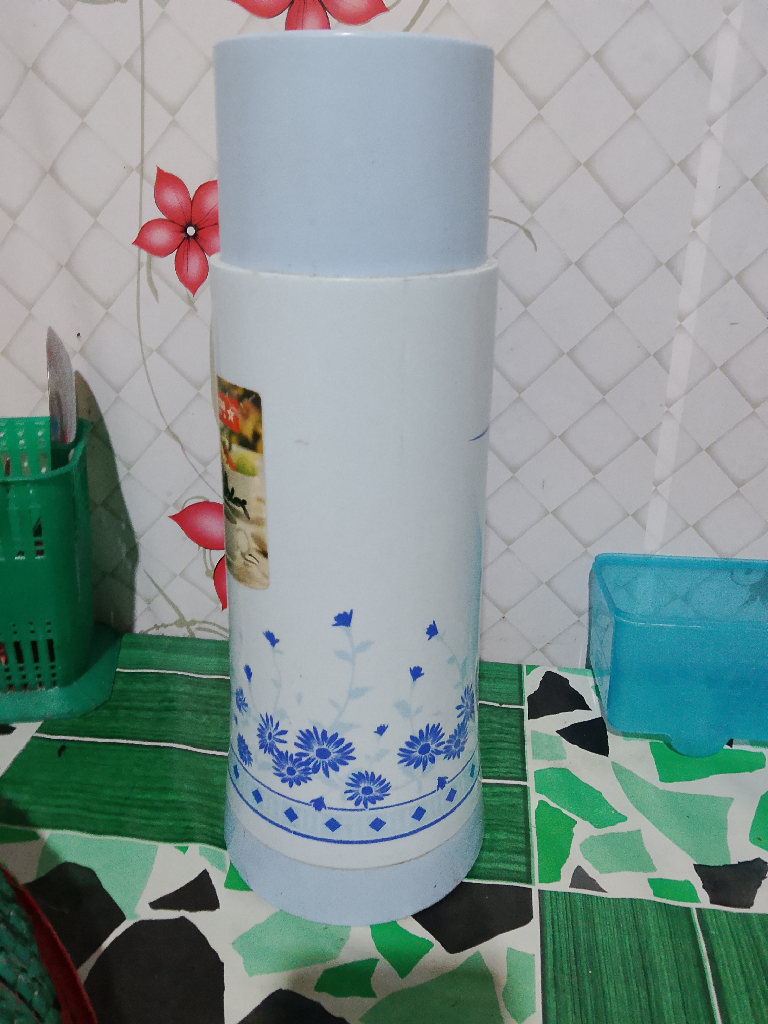 A light blue and white cylindrical thermos with a floral pattern is placed on a green and white patterned surface. The thermos has a lighter blue cap. To the left of the thermos is a green utensil holder with a knife inside. To the right of the thermos is a light blue plastic container. The background consists of a tiled wall with a red floral pattern. The scene appears to be an indoor setting, possibly a kitchen counter in Merbopuntong, Indonesia.