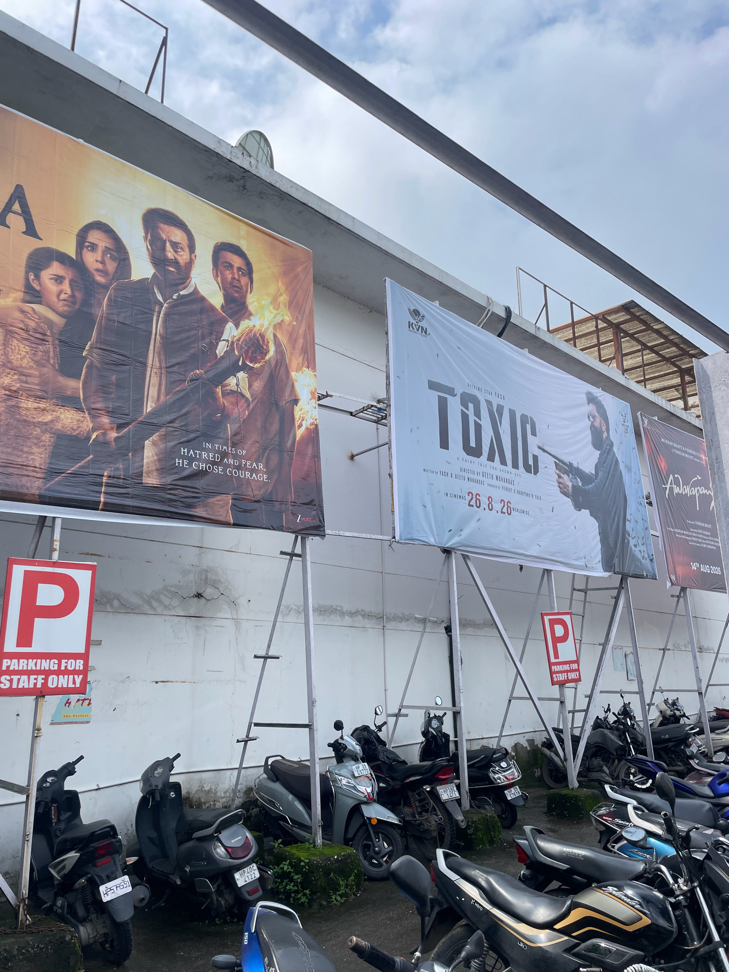 The image captures an outdoor scene in Dharamshala, India, featuring several movie posters displayed on a white wall. In the foreground, a row of parked motorcycles and scooters is visible. Two red and white "P PARKING FOR STAFF ONLY" signs are prominently displayed, one on the left and another further back on the right. The movie posters are large banners mounted on metal scaffolding. The poster on the left appears to be for a film with the title starting with "A", featuring several actors, including a prominent male figure holding a burning torch. Text on this poster reads "IN TIMES OF HATRED AND FEAR, HE CHOSE COURAGE." To the right of this, a large poster for the film "TOXIC" is displayed, with the title in bold, grey lettering and an image of a man holding a gun. Below the title, it indicates "IN CINEMAS 26.8.26". Further to the right, a portion of another movie poster is visible, with the title "ADARAPANT" in red and black lettering. The sky above is cloudy. The overall environment suggests an outdoor area, possibly near a cinema or event venue.